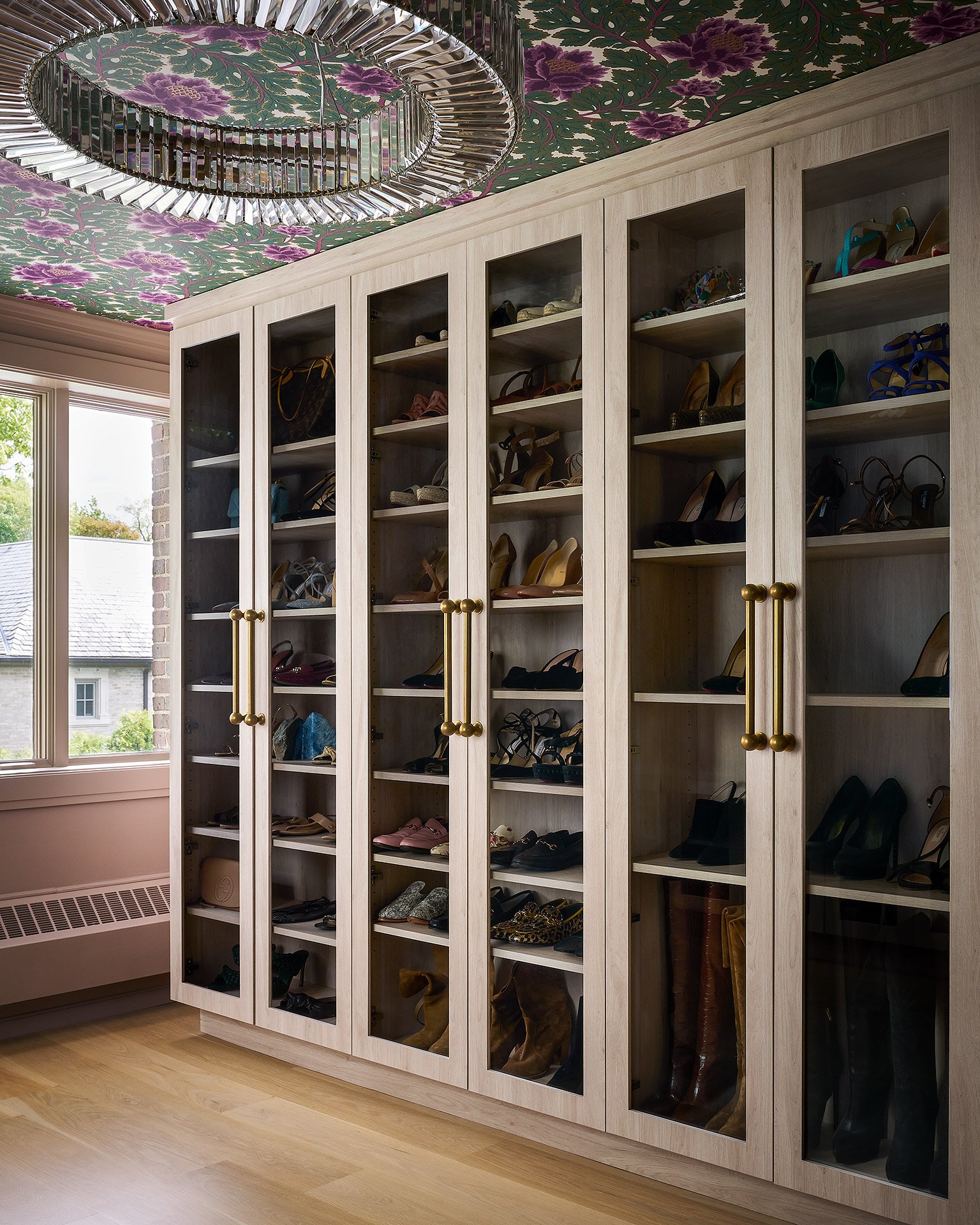 A wooden shoe cabinet with glass doors filled with various high-heeled shoes, situated near a window with a floral wallpaper ceiling and a metallic chandelier.