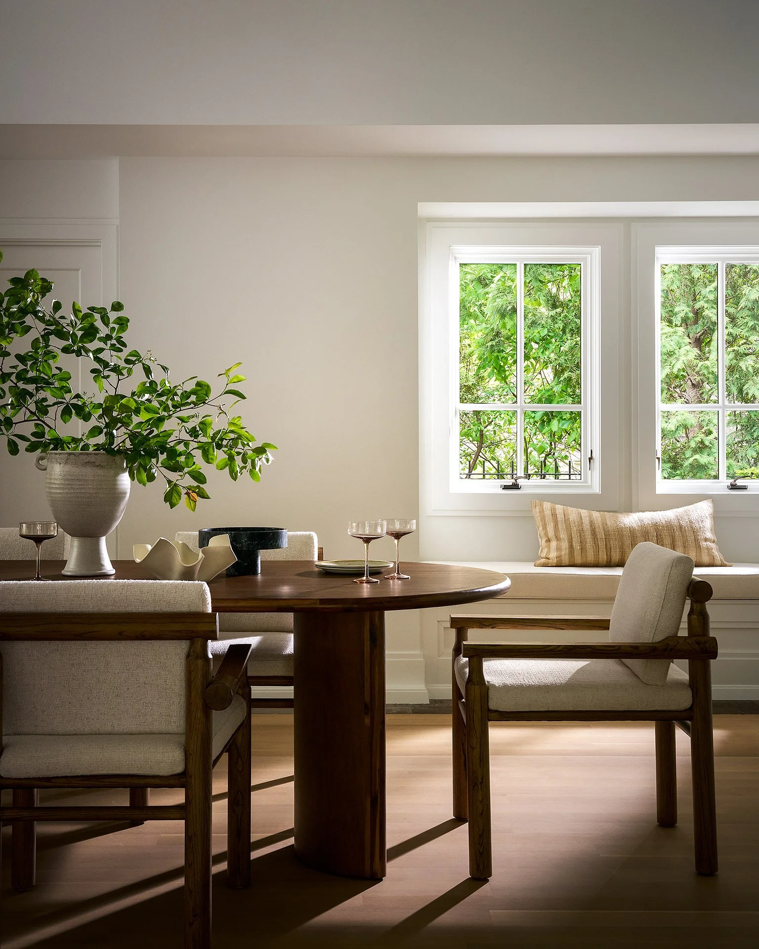 A cozy dining area illuminated by natural light from two large windows with views of green trees. The space features a wooden table, upholstered chairs, and a side bench with a striped pillow, decorated with a large green plant in a white pot and som