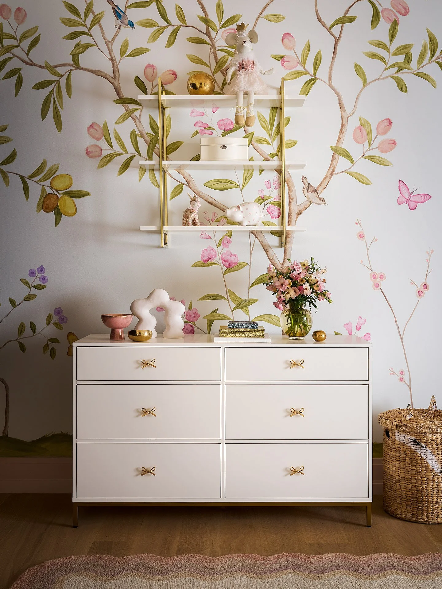 White dresser with gold bows on drawer pulls, decorated with various decor items and a floral wallpaper background.