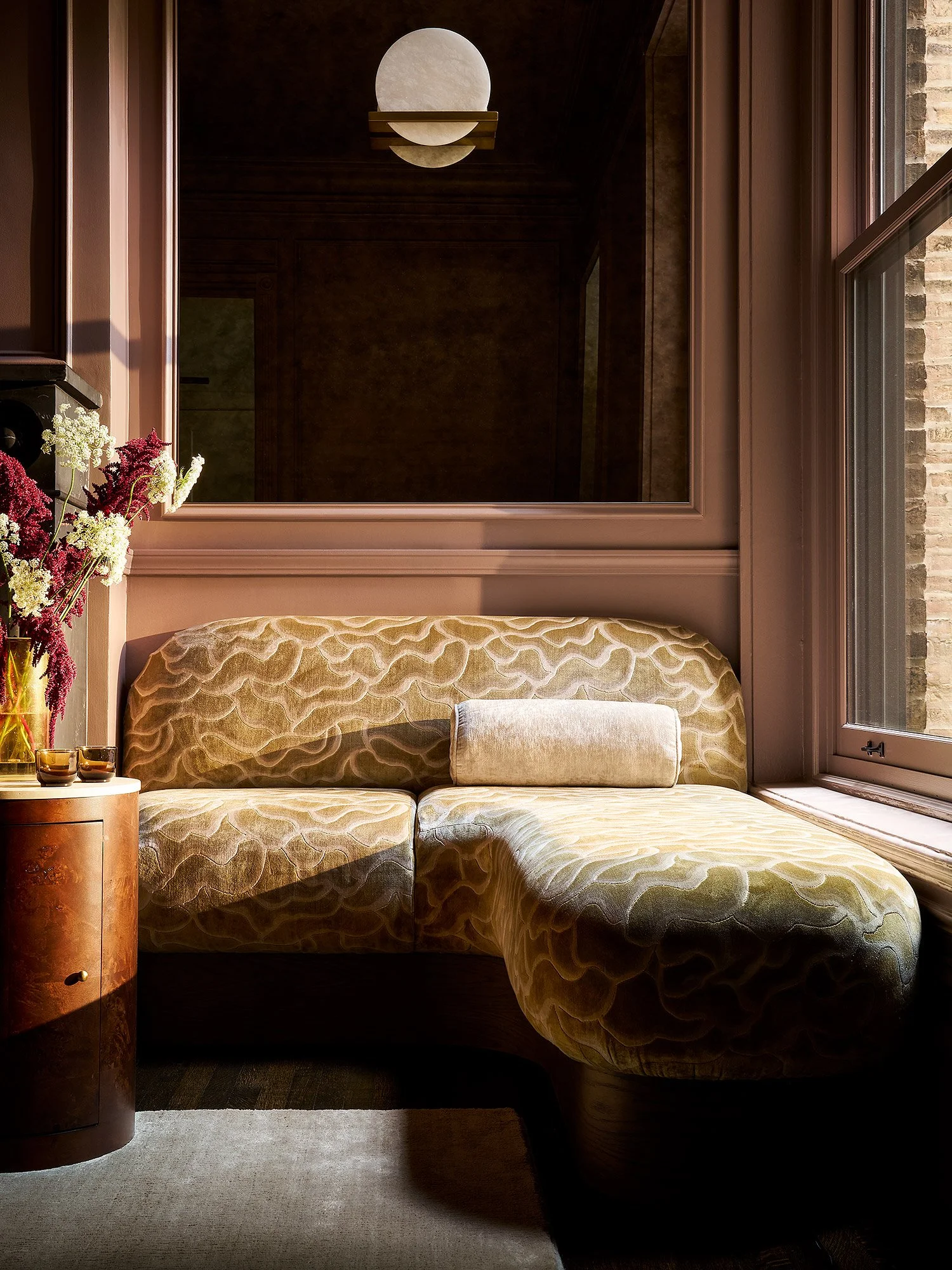 A cozy nook with a beige velvet curved sectional sofa with a matching bolster pillow, a small wooden side table holding a vase of fresh flowers and two candles, a large window letting in natural light, and a modern wall-mounted light fixture against 