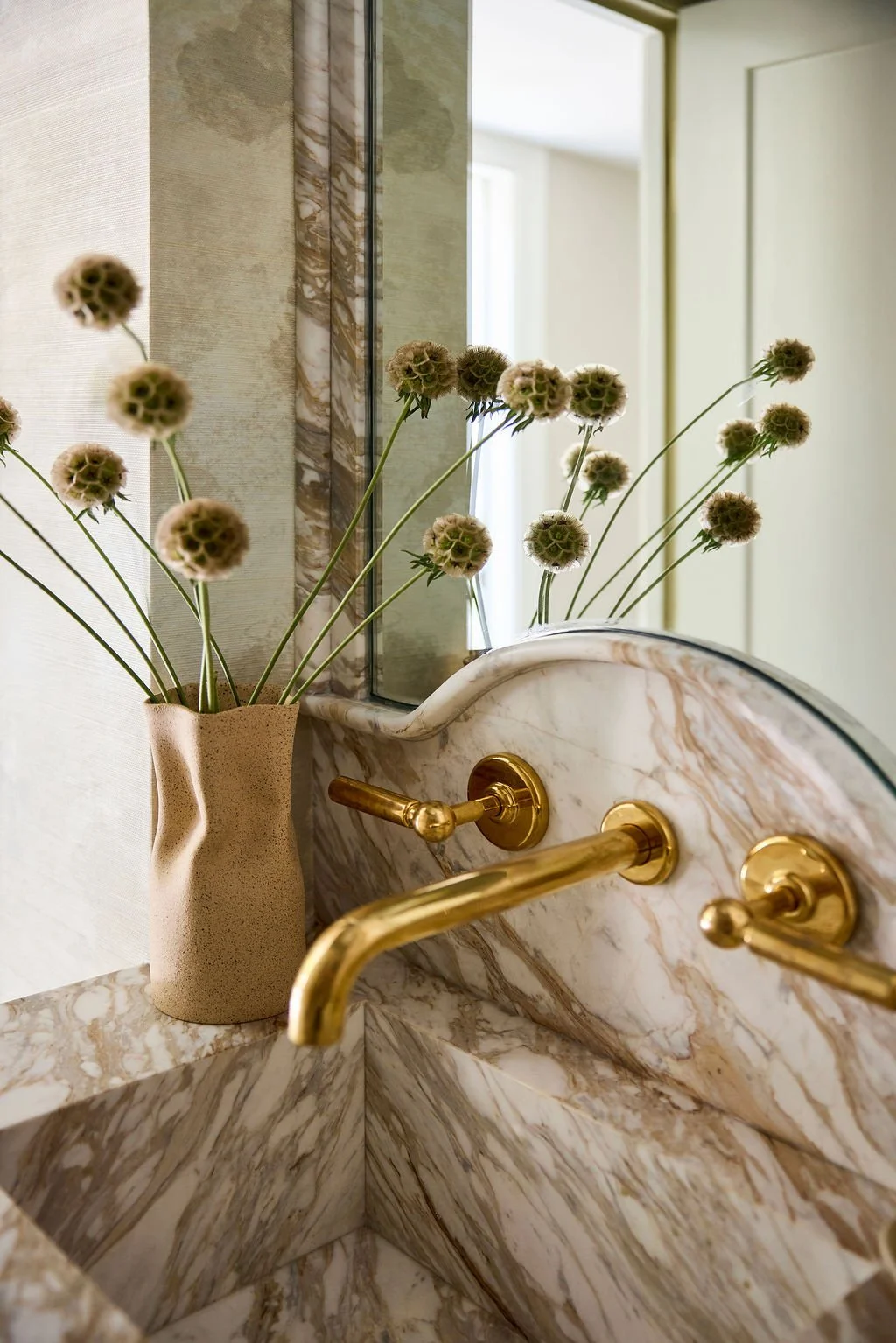 A marble sink with gold fixtures and a mirror, decorated with a beige vase holding dried flowers, in a bathroom with light-colored walls.