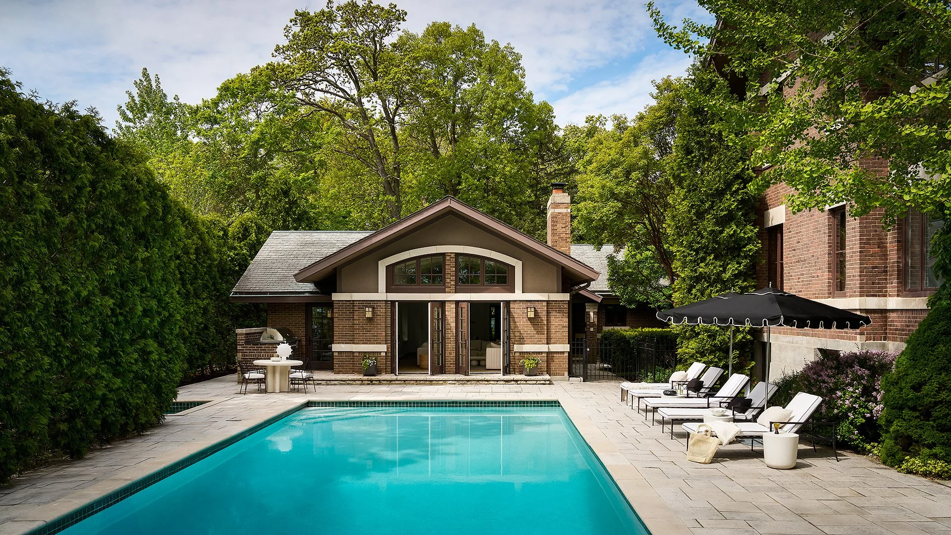 Backyard with swimming pool, patio furniture, and a house with brick walls and large windows, surrounded by lush green trees and shrubs.