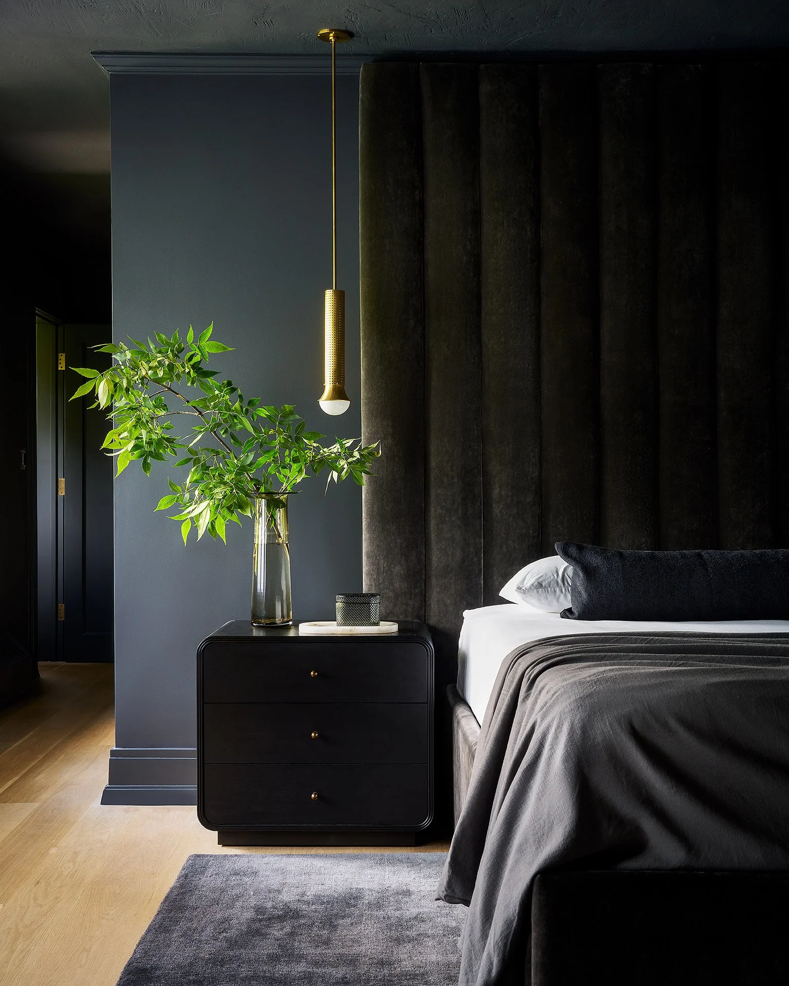 A bedroom with a dark color theme, featuring a black nightstand with gold knobs, a tall green plant in a glass vase, a modern gold pendant light, a dark upholstered headboard, and a bed with white sheets and gray bedding.