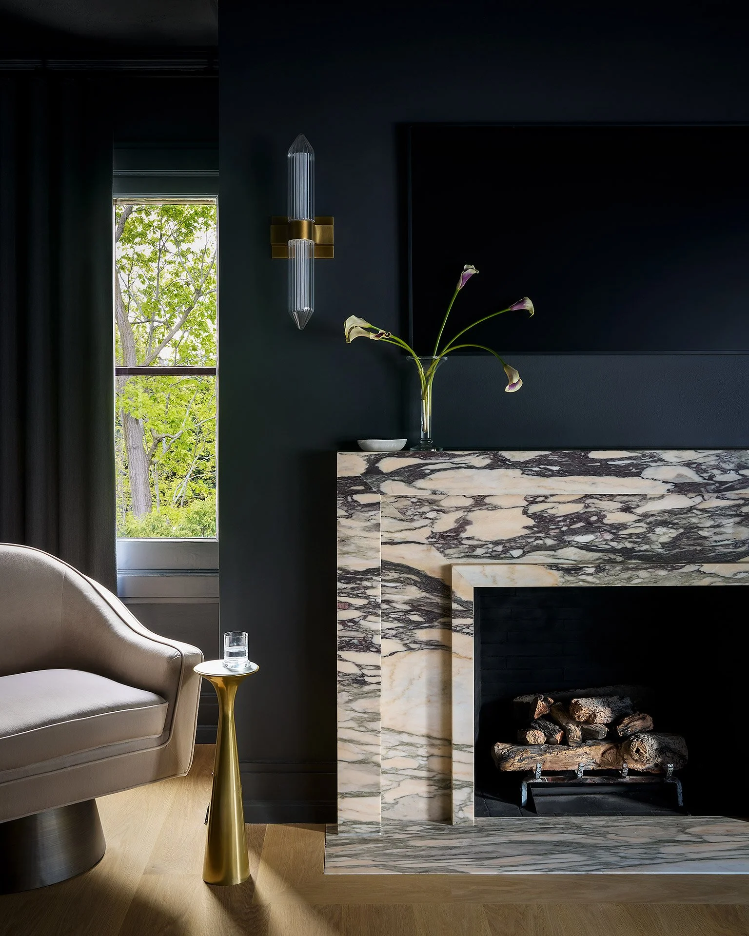 A living room corner with a white armchair, a gold side table with a glass of water, a dark wall with a modern vertical wall sconce, a black TV, green trees outside the window, and a marble fireplace with logs.