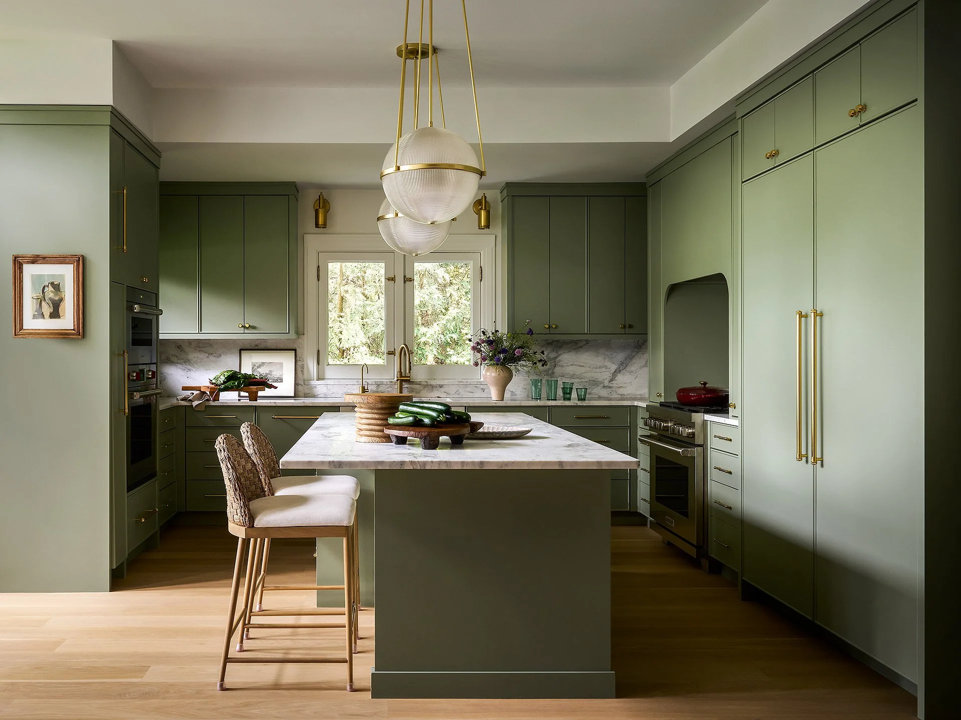 Green kitchen with marble countertop island, gold hardware, and two chairs at the island. Large window behind the sink, pendant lights overhead, and various kitchen items on the counter.