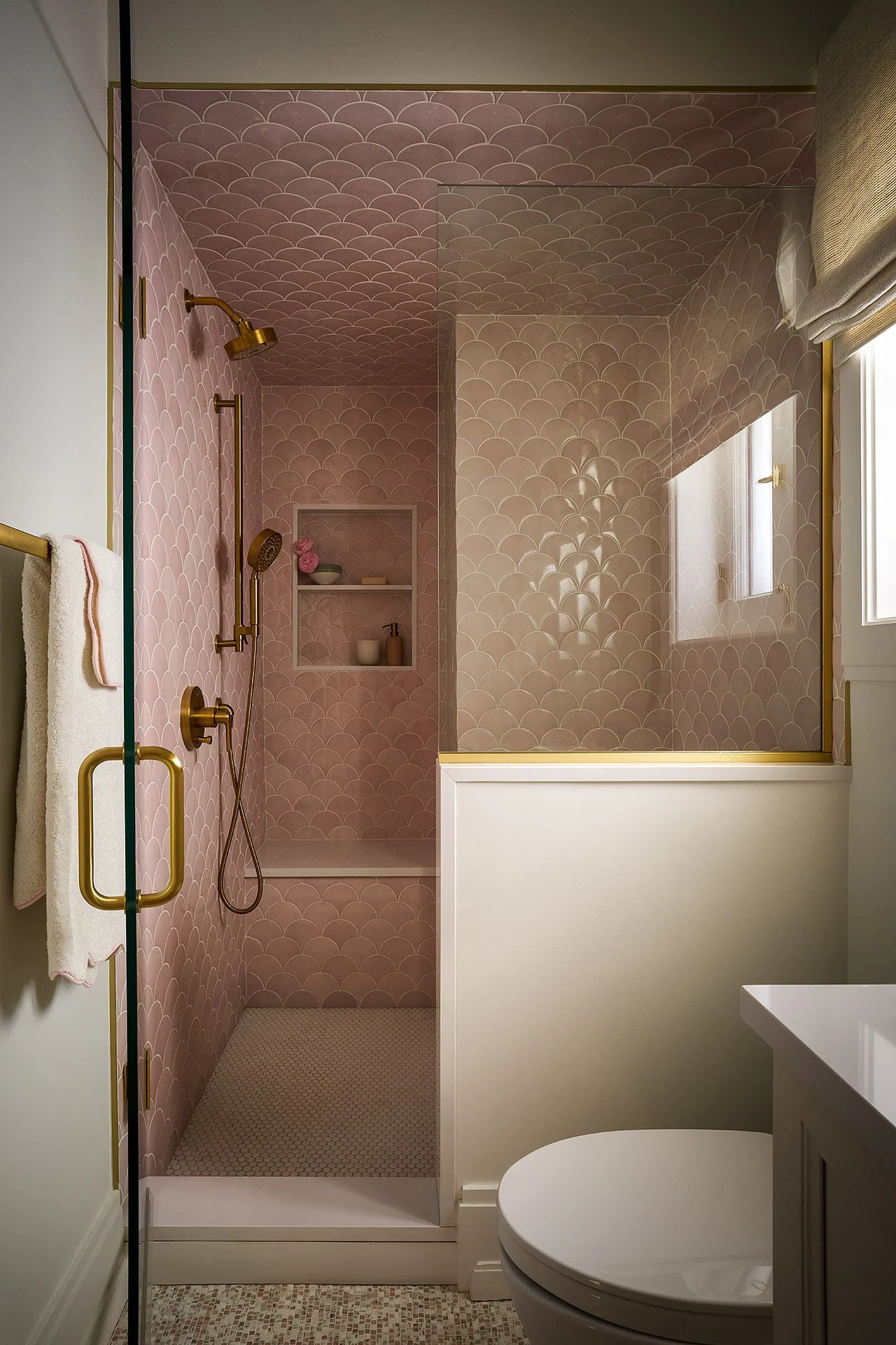 Interior of a bathroom with a pink scallop tile shower, gold fixtures, a built-in shelf with pink flowers, and a white toilet beside a vanity with a white countertop.