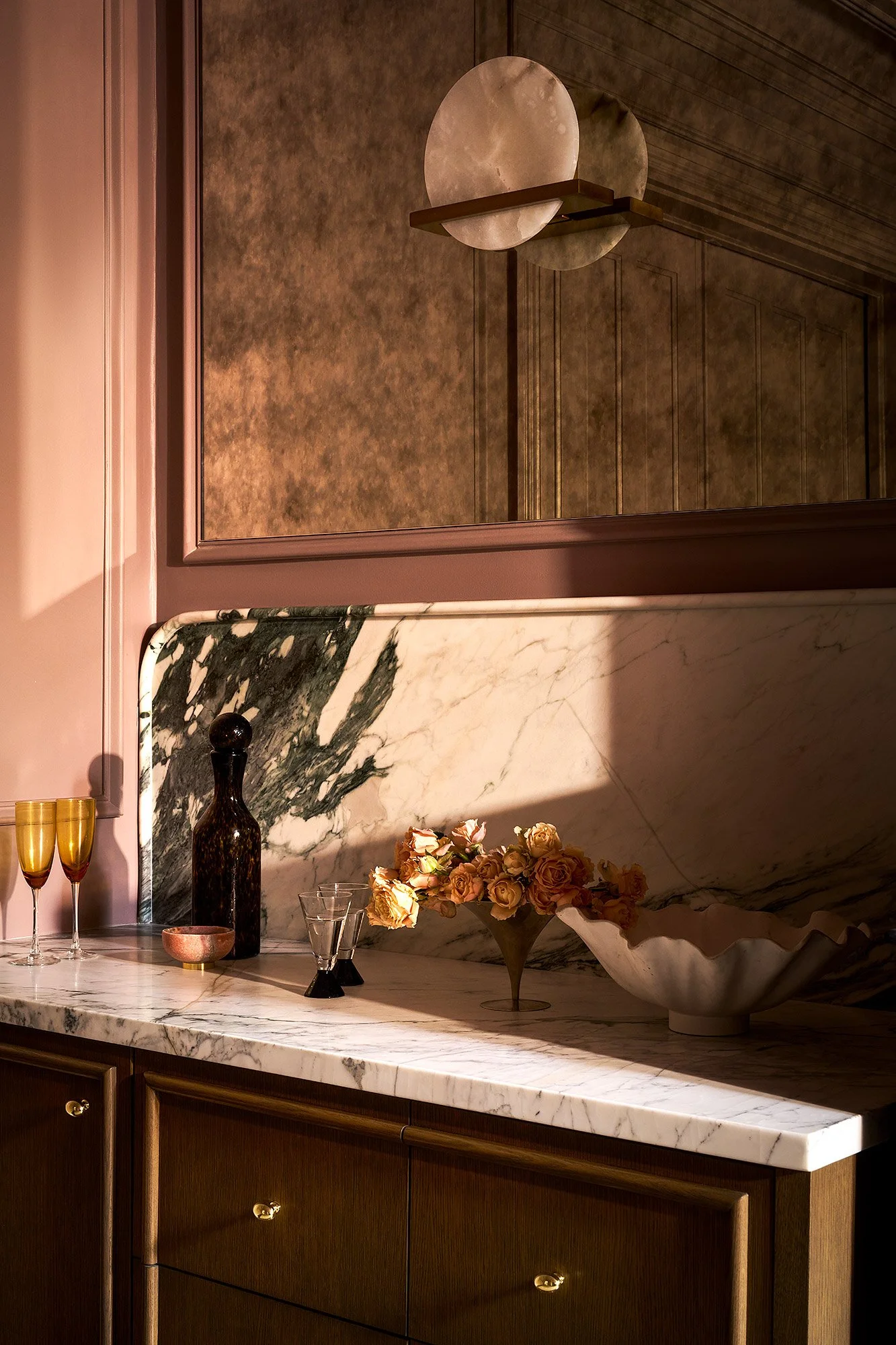 A marble countertop with a flower arrangement in a vase, two glasses, and two gold-colored glasses. A black vase and a pink bowl are also on the counter. In the mirror, a section of textured wall and a wooden cabinet are visible.