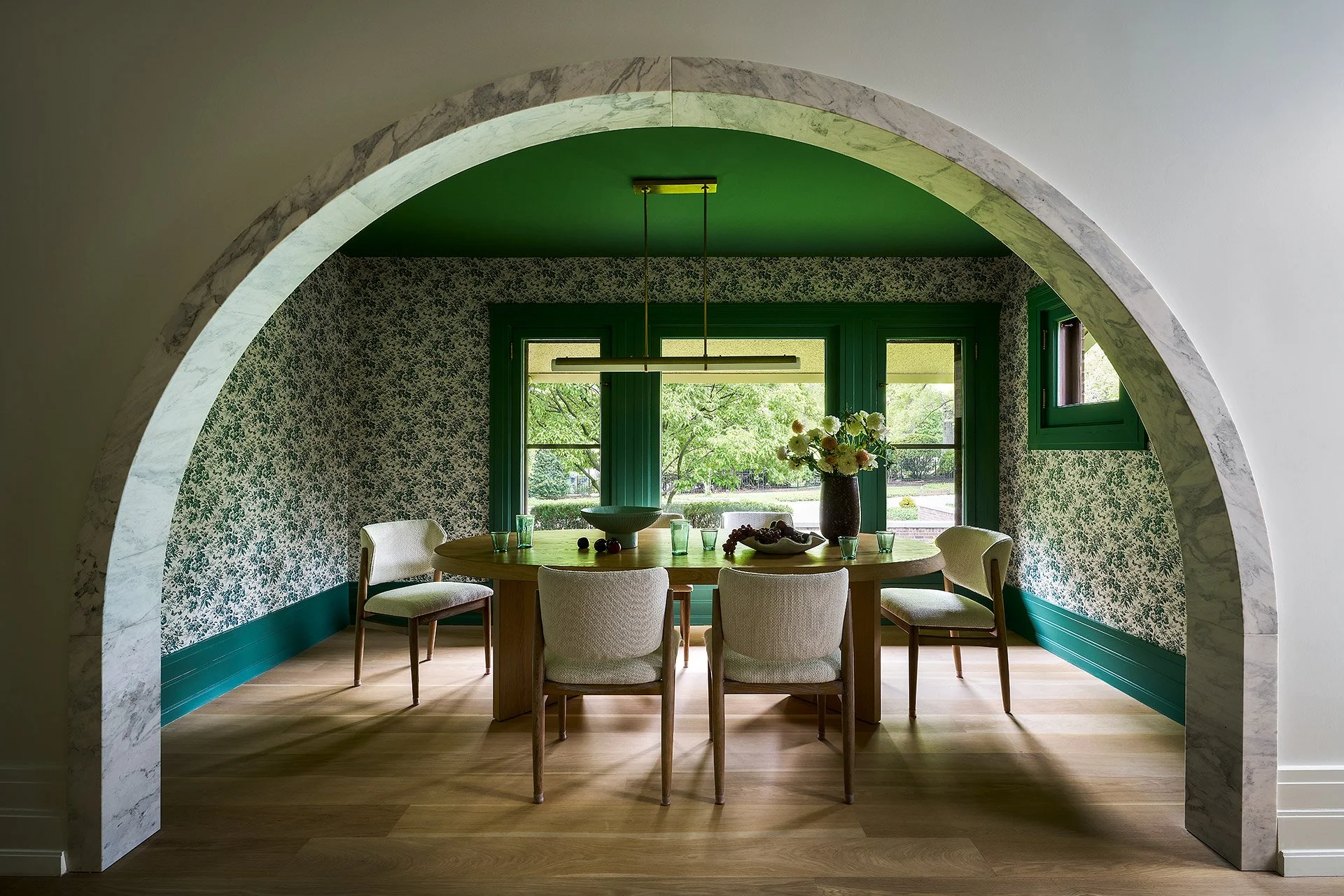 A dining room with a circular wooden table surrounded by beige upholstered chairs. There is a vase of white flowers and a fruit bowl on the table. The room has a green ceiling and window frames, with patterned wallpaper, and is viewed through a marbl