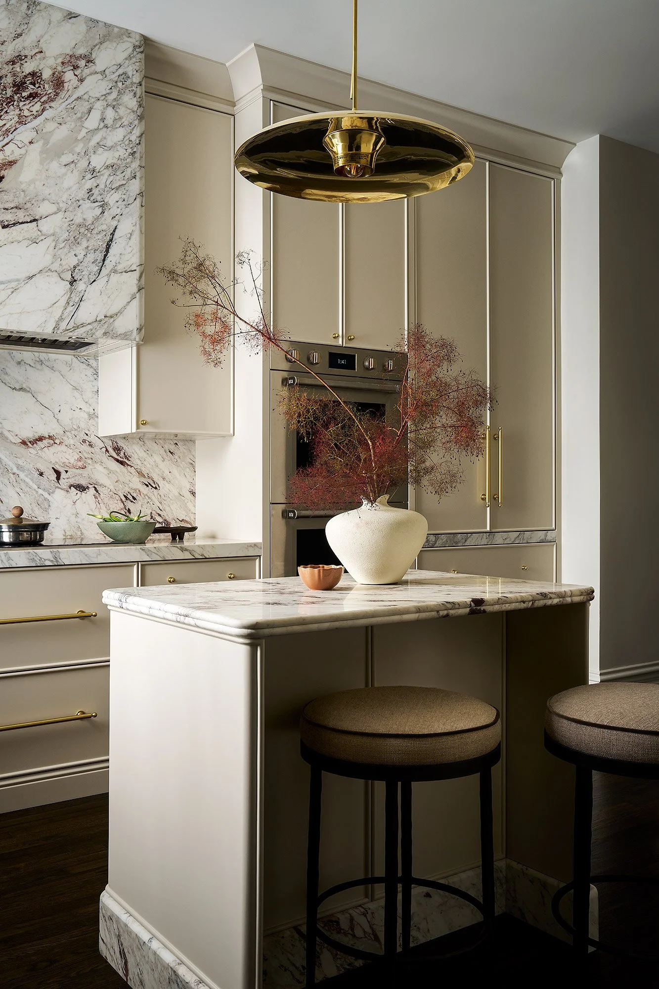 Modern kitchen with beige cabinets, marble countertops, a large marble backsplash, and a marble kitchen island. Two brown bar stools are positioned at the island. A tall beige vase with dried branches sits on the island, and a small bowl with condime