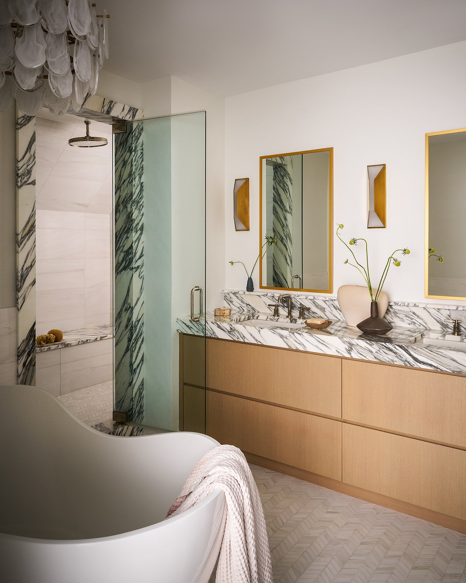Modern bathroom with a large bathtub, vanity with marble countertop, two mirrors with gold frames, a white vase with flowers, and a glass shower stall with marble accents.