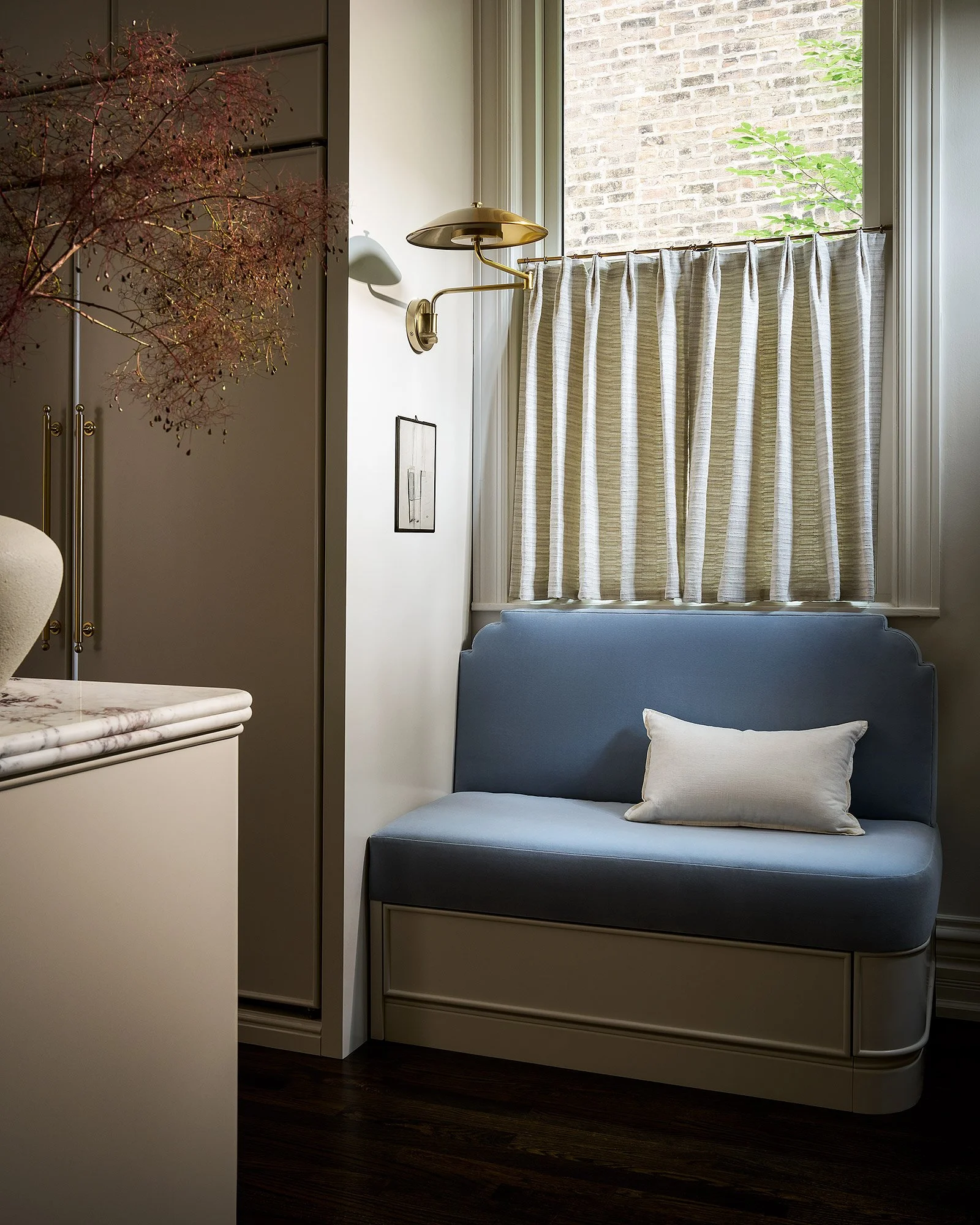 A cozy corner with a blue upholstered bench, a white pillow, a brass wall-mounted reading light, a window with striped curtains, and a brick wall outside.