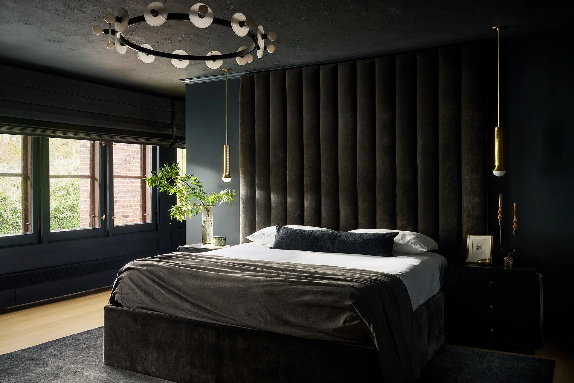 Modern bedroom with dark walls, large bed with white and dark pillows, natural light from windows, decorative pendant lights, green plant, framed artwork, and minimal decor.