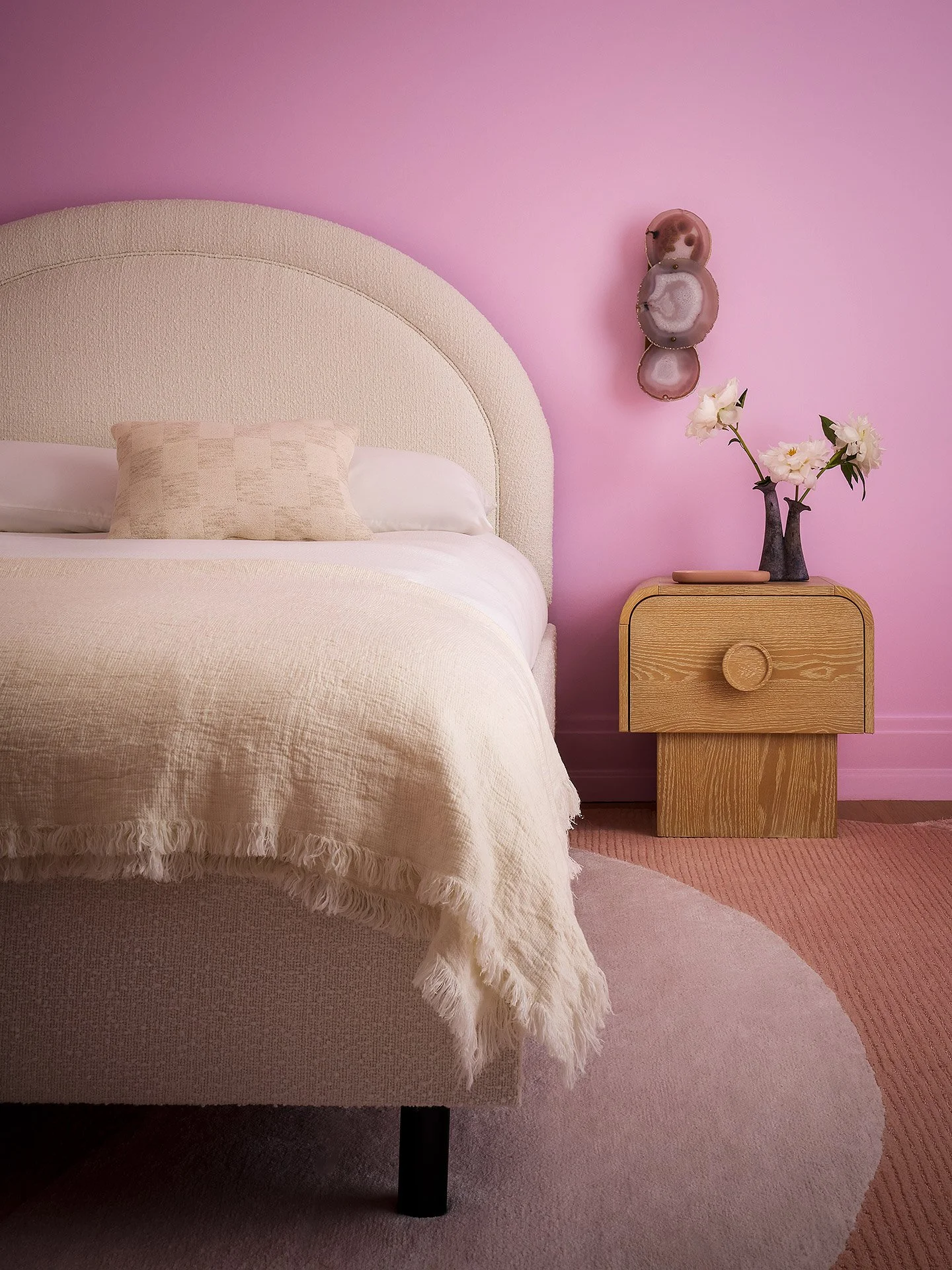 A bedroom with a bed, beige headboard, white bedding, and a beige pillow. To the right, a wooden nightstand with a black vase holding white flowers, on pink walls with decorative agate or geode wall art.