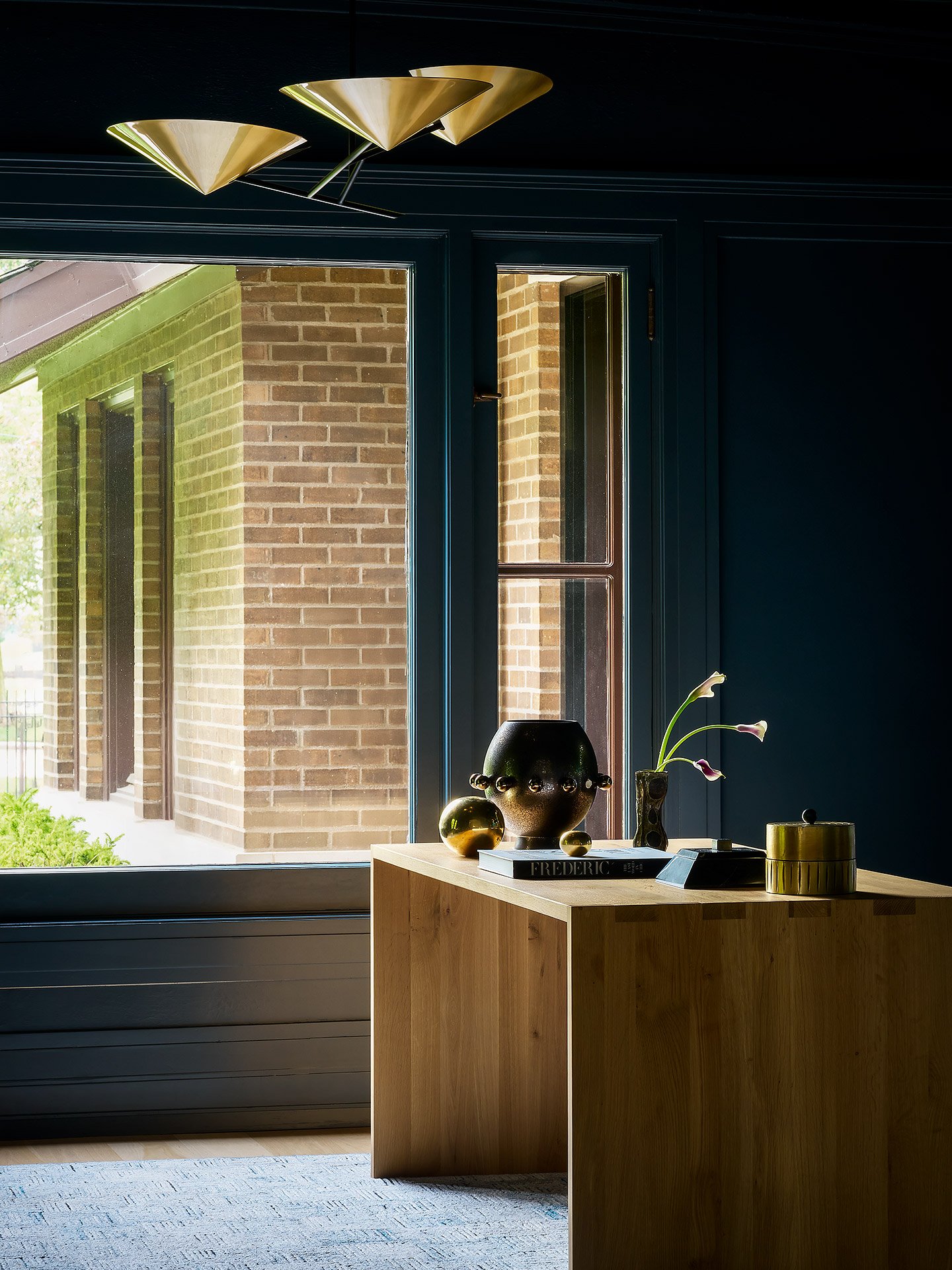 Interior view showing a wooden table with decorative items and a large window revealing the exterior brick wall of a building and some greenery.