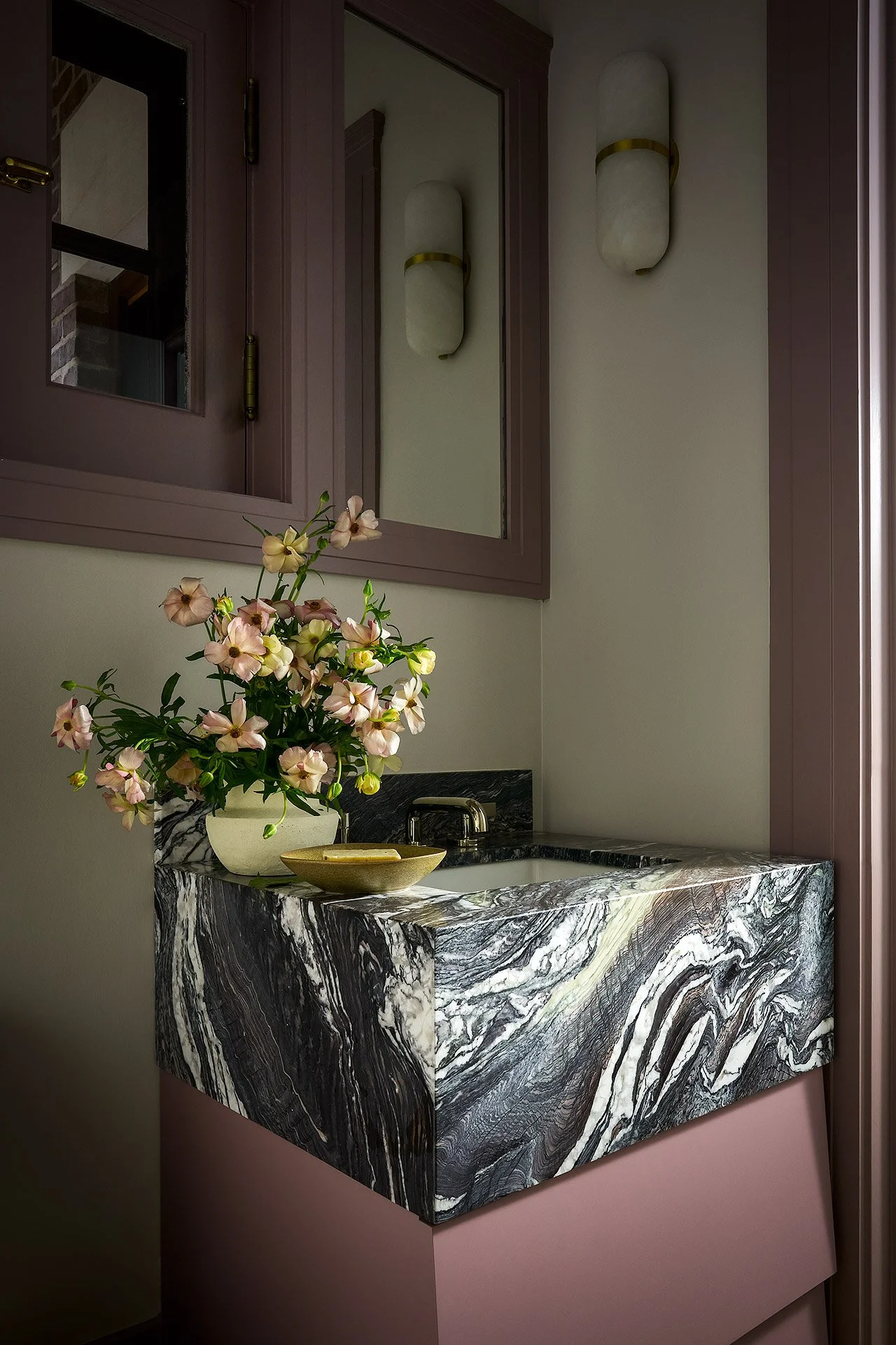 A bathroom with a black and white marble countertop sink, a vase with pink and cream flowers, a mirror, and wall-mounted lights.