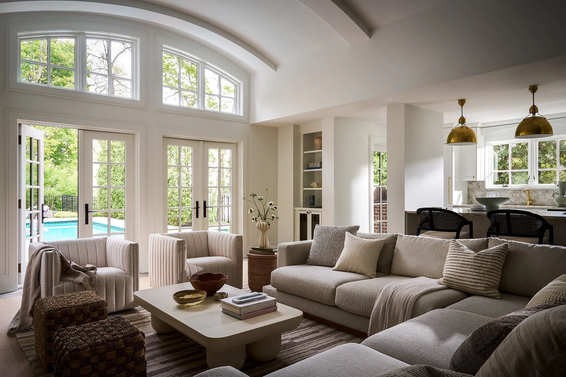 Bright living room with large windows and glass doors leading to a backyard pool, beige sofas, striped chairs, and a white coffee table with bowls and books.