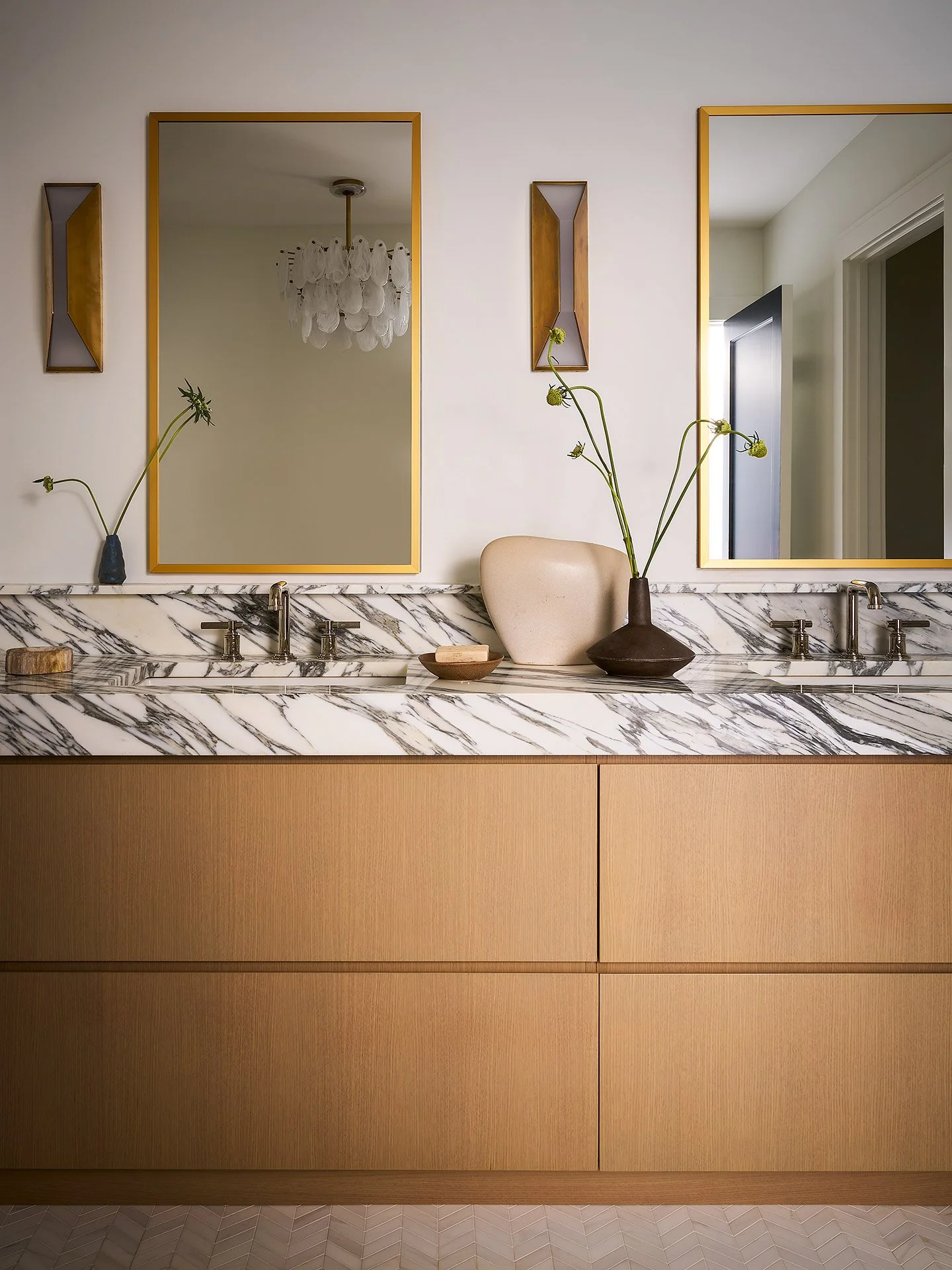 A bathroom vanity with a marble countertop, two sinks, and two large mirrors with gold frames. Decor includes vases with flowers, a soap dish, and small wall decorations. The cabinetry is wooden with a warm finish.
