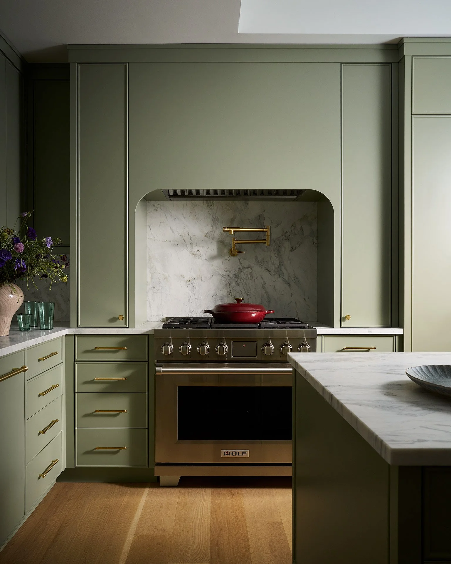 A kitchen with green cabinetry, a marble backsplash, a red pot on a stainless steel stove, a marble countertop, and a vase of flowers on the left.