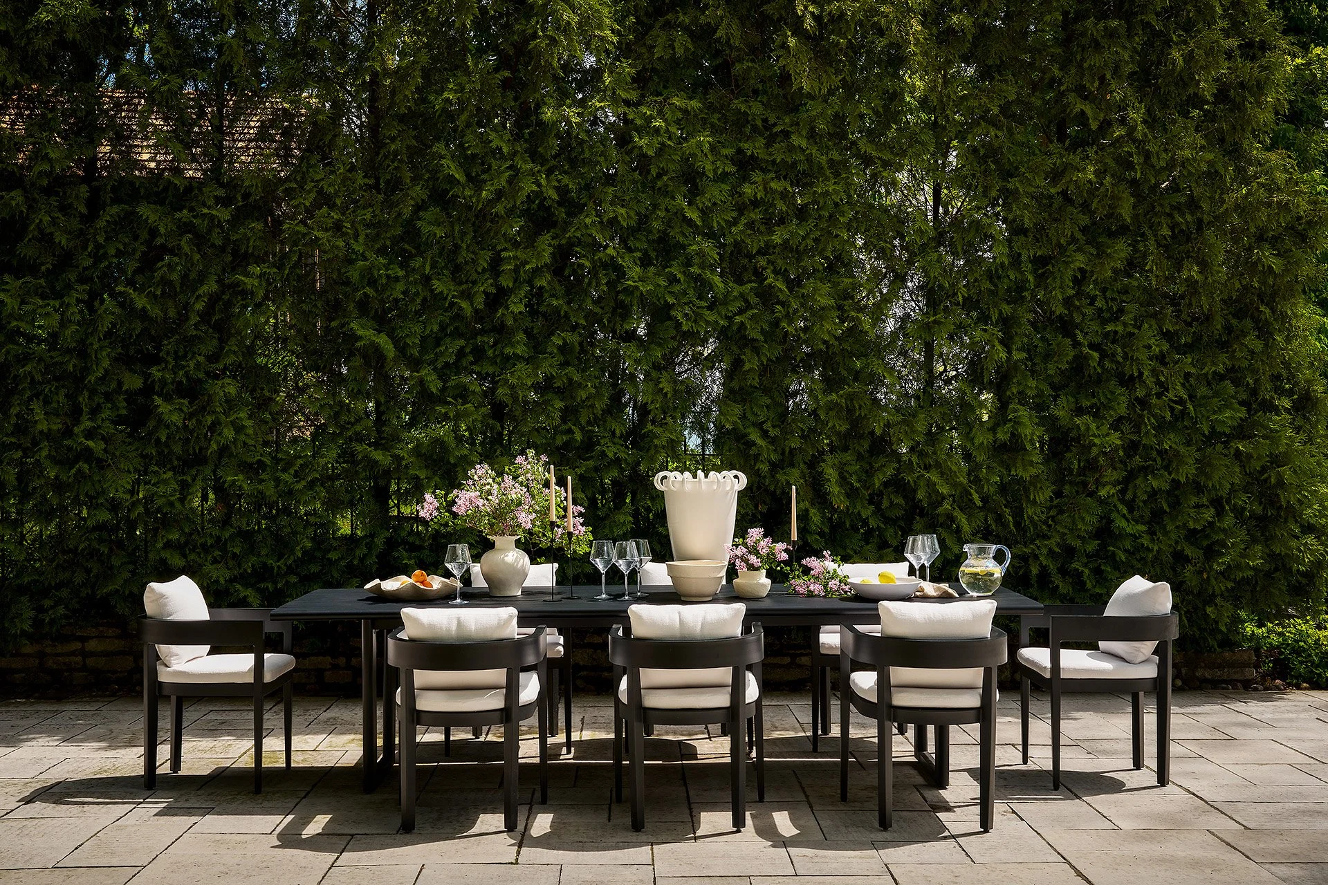 Outdoor dining table with white chairs, set with wine glasses, vases of pink flowers, a pitcher of water, and a large white decorative vase, against a backdrop of tall green trees.