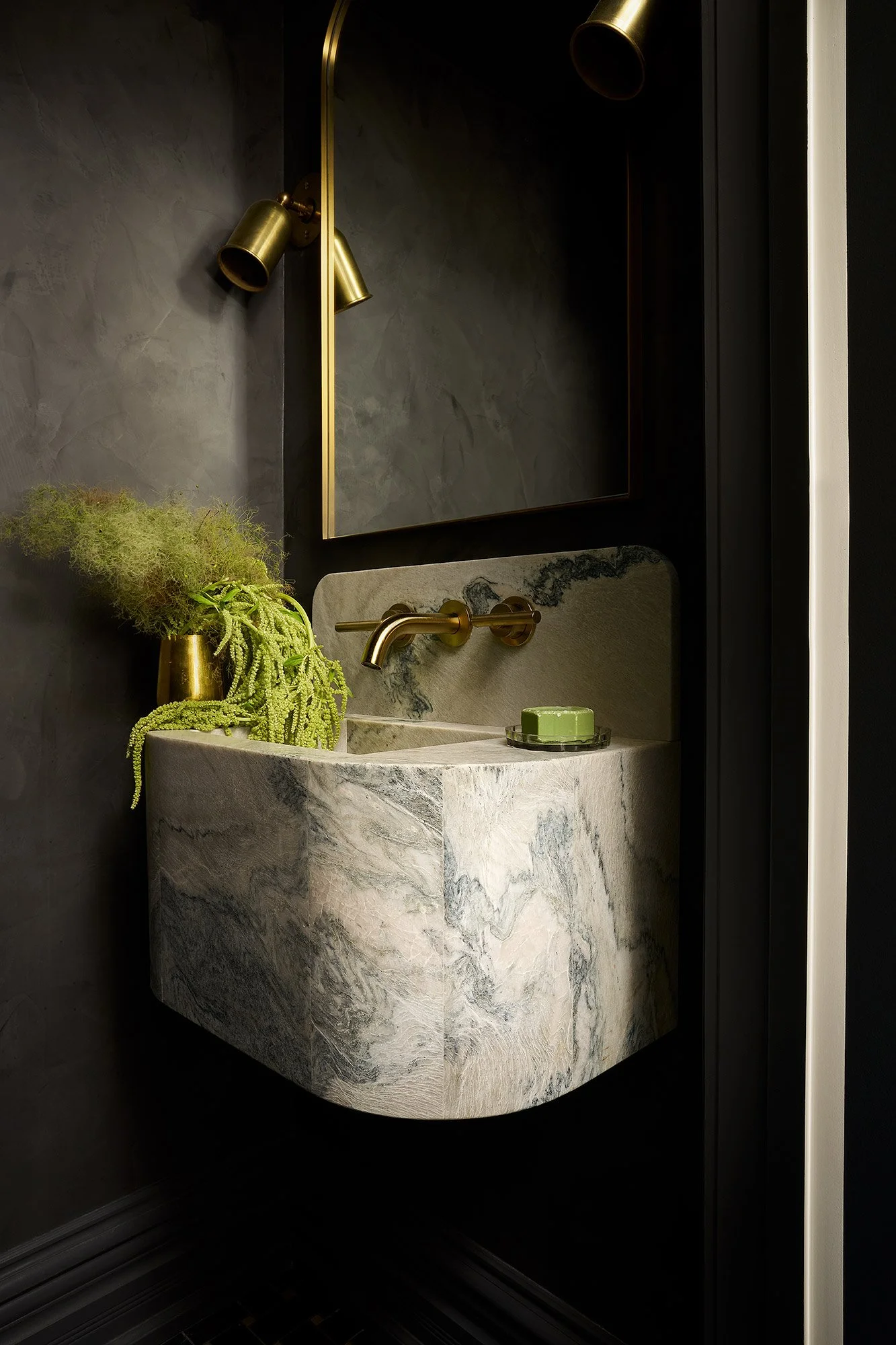 A wall-mounted marble sink with gold fixtures, a mirror with a gold frame, and a small green soap dish. There is a black wall with a plant in a gold vase next to the sink.