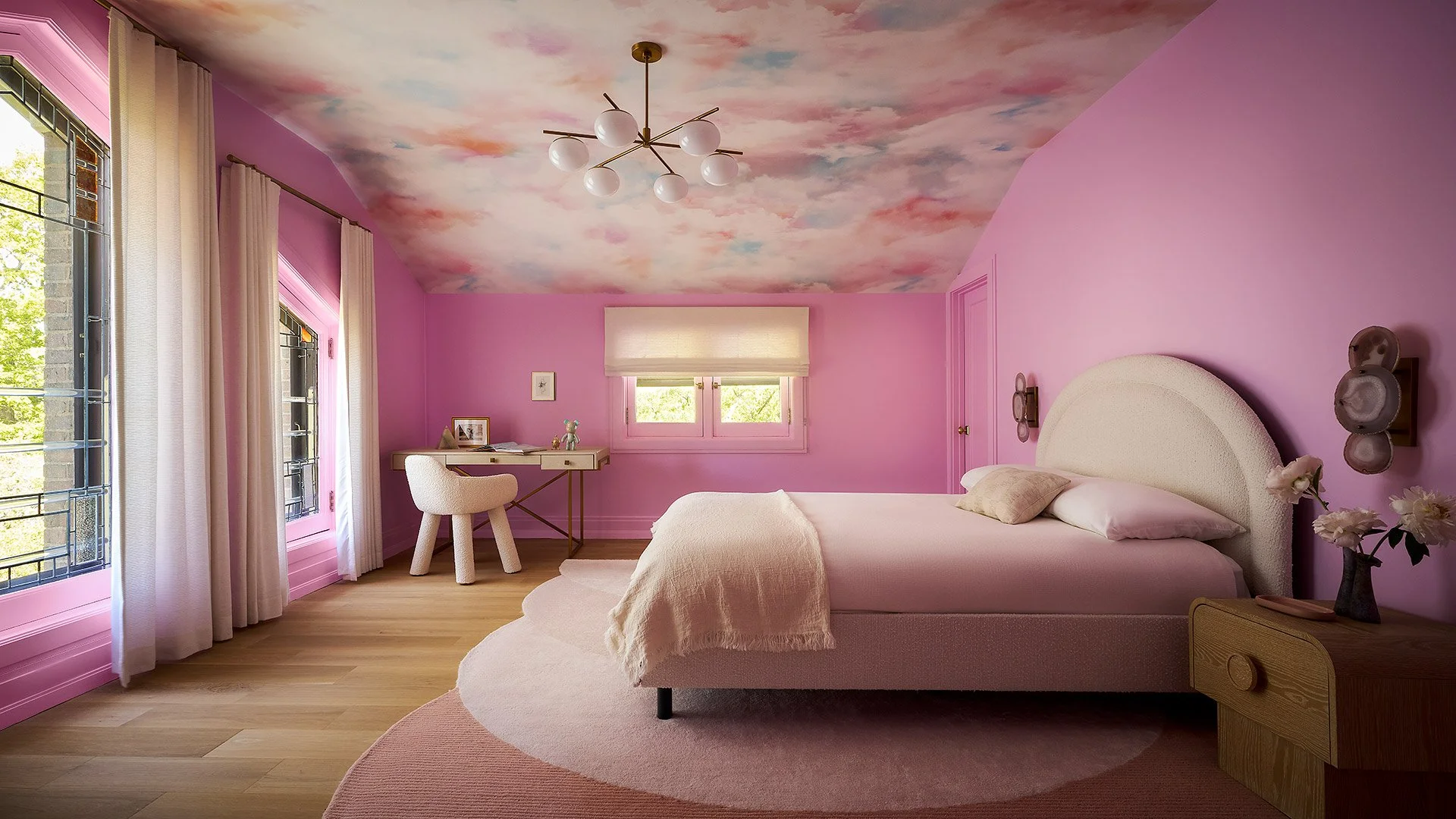 A bedroom with pink walls and cloud-painted ceiling, featuring a large bed with white bedding and a rounded headboard, a nightstand with a vase of flowers, windows with white curtains, a small write desk with a chair, and modern light fixtures on the
