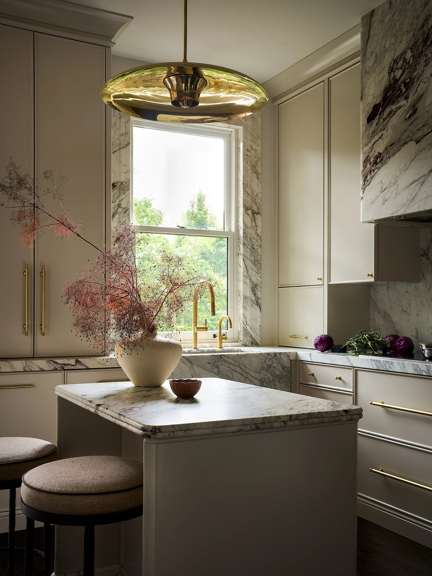 A modern kitchen with white cabinets, marble countertops, and a marble backsplash. A window lets in natural light. A large brass pendant light hangs from the ceiling. A white vase with pink flowers and a small bowl sit on the kitchen island, with two