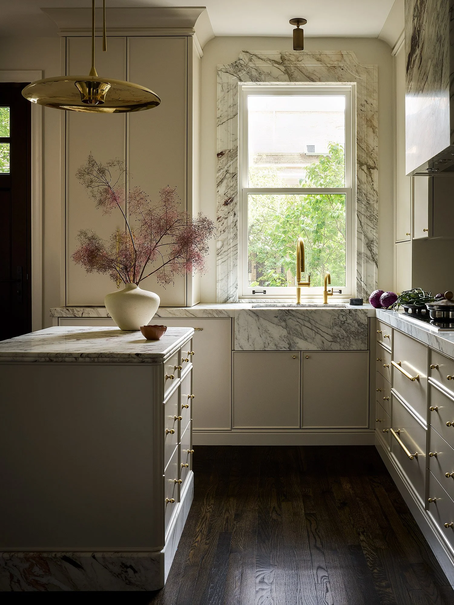 Modern kitchen with white cabinetry, marble countertops, a window with marble trim, and dark hardwood floors. A vase with pink dried flowers is on the counter.