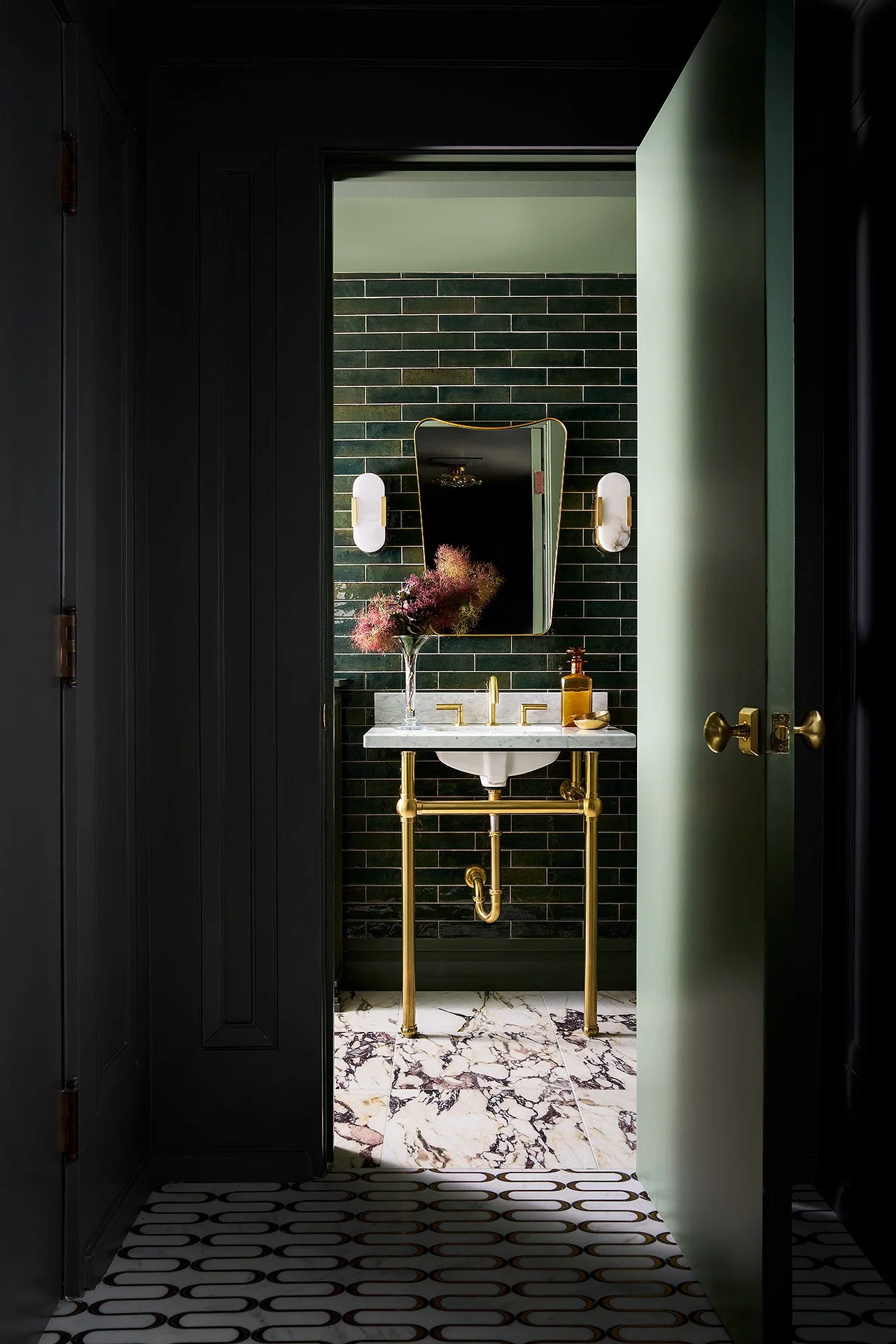 A small bathroom with black tiled wall, white marble sink with gold fixtures, green walls, and a mirror with wall sconces. There is a black door partially open, revealing a patterned tile floor with black, white, and gold accents.