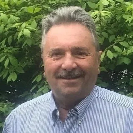 Headshot of Michael Bruno standing outdoors in front of green foliage.