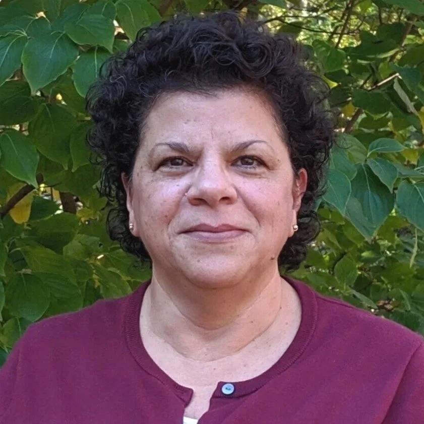 Headshot of Debbie Roberts standing outdoors in front of green foliage.