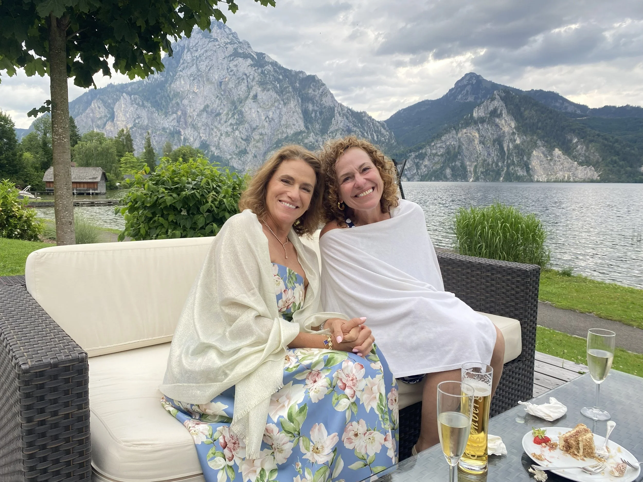 Twin sisters and business partners Tammy and Tracy smiling together lakeside in the Austrian Alps, reflecting the personal bond behind their team alignment and leadership consulting work