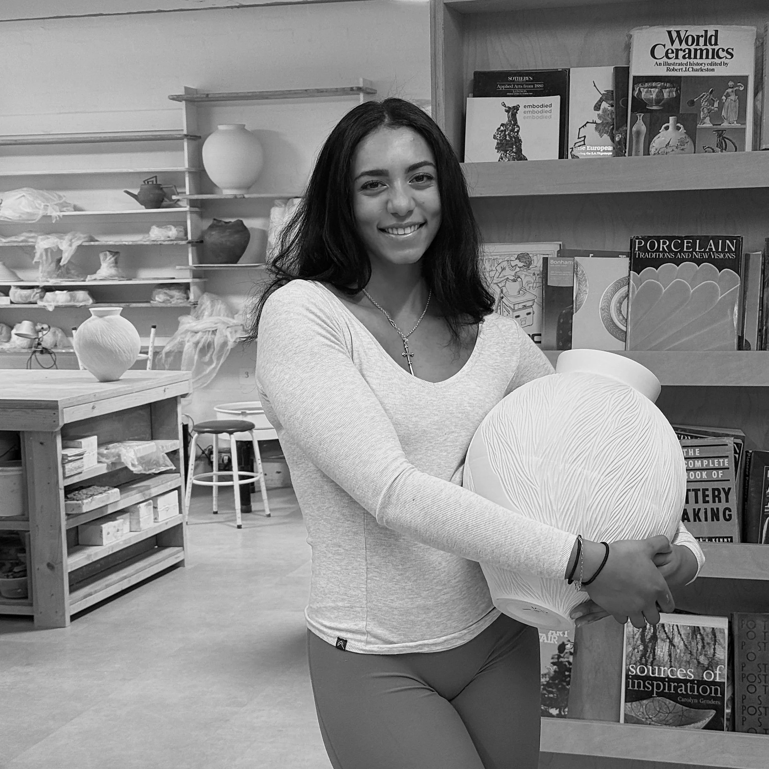 Young, talented woman who is a pottery teacher from the USA smiling and holding a large ceramic vase in a ceramics or pottery studio, with shelves of books and ceramic supplies in the background.