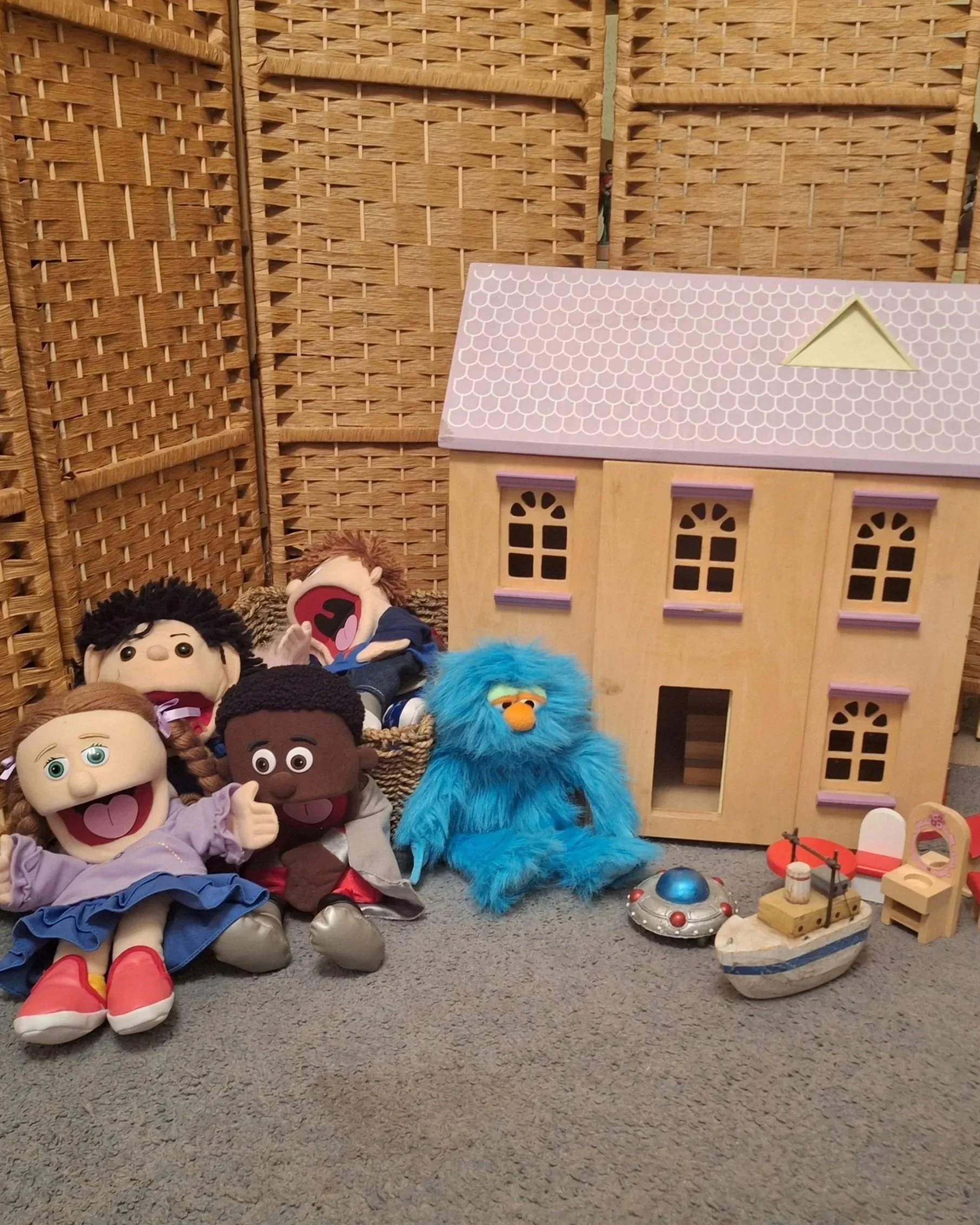 A collection of different puppets sat on carpet next to a pink and brown dolls house with dolls furniture and a wooden boat in front.