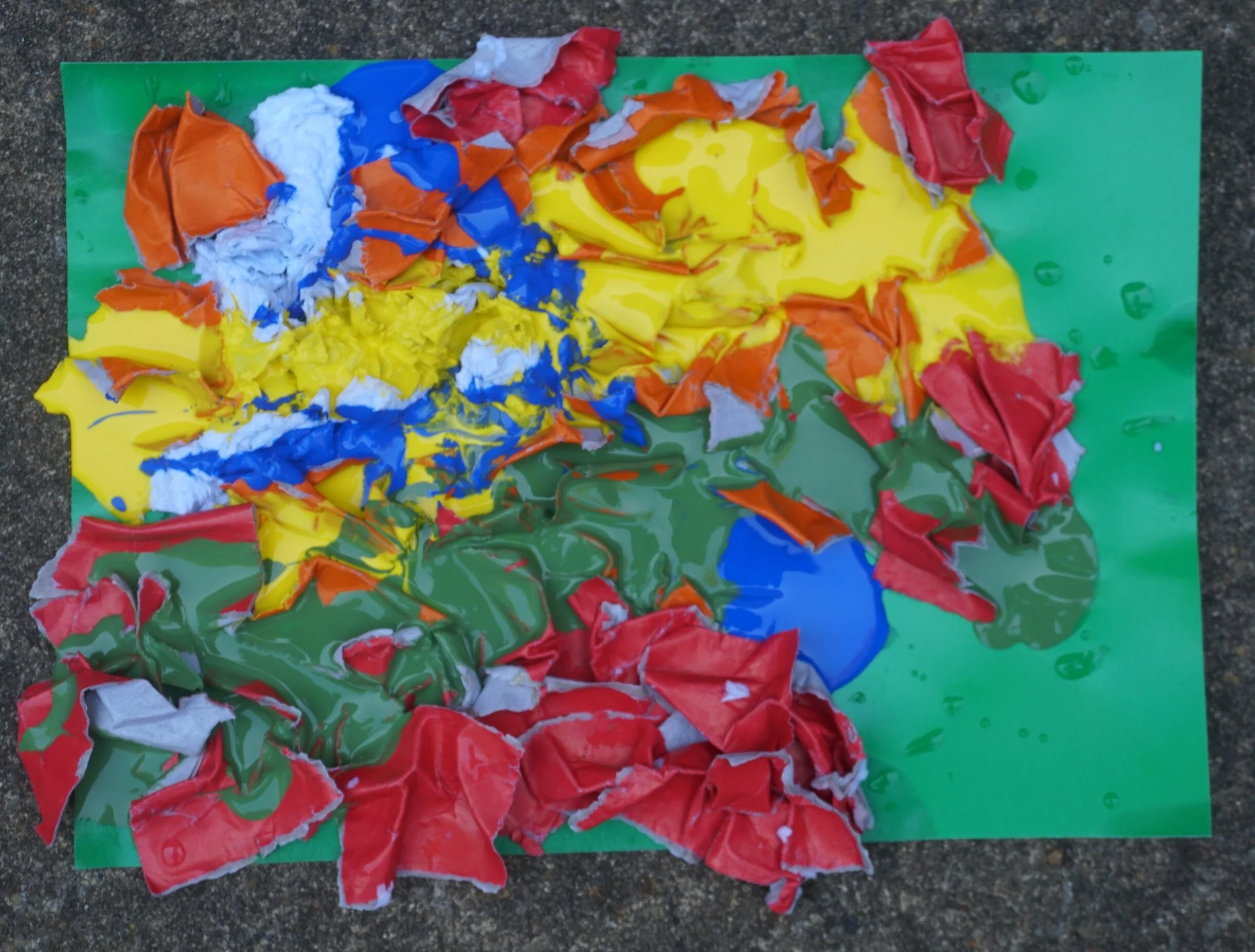 Green piece of card with red, blue, green and yellow paint and paper stuck to it randomly.