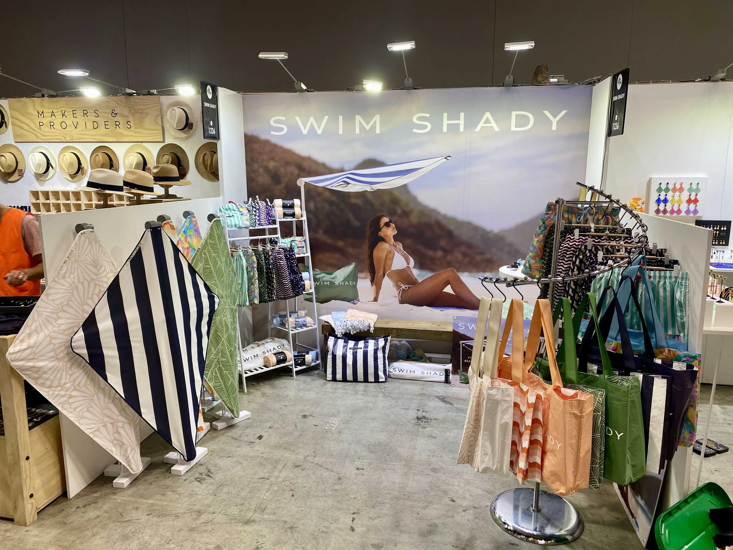 Swim Shady trade show booth featuring a large full-size beach lifestyle photo print of a woman in a bikini lounging by the sea, surrounded by displays of striped beach bags, patterned towels, and sun umbrellas.