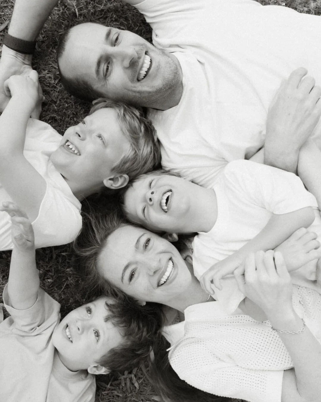 A group of six people, two adults and four children, lying on the grass and smiling, captured in a black-and-white photo.