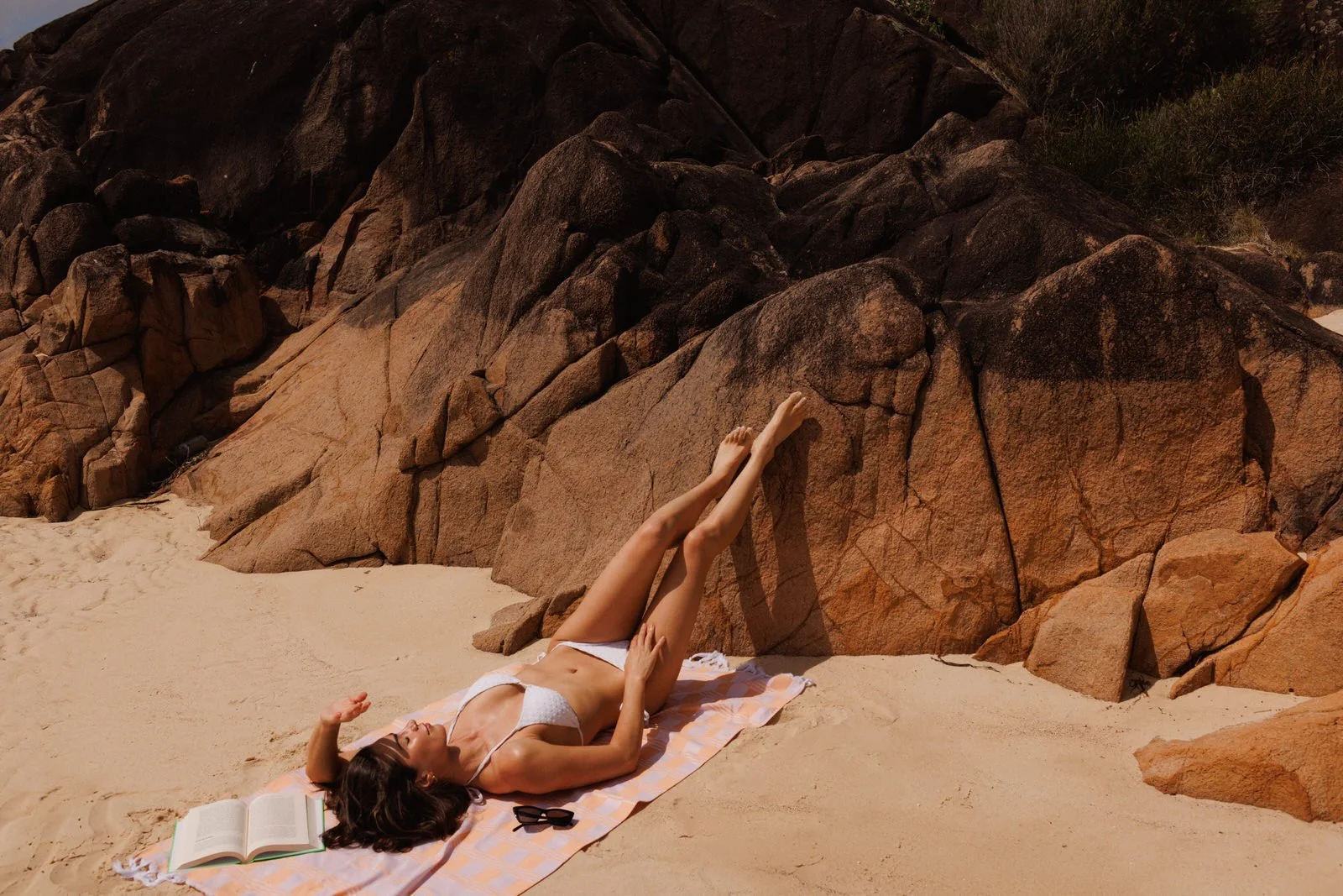 Woman in a white bikini lying on a beach towel by rocks, relaxing with a book nearby.