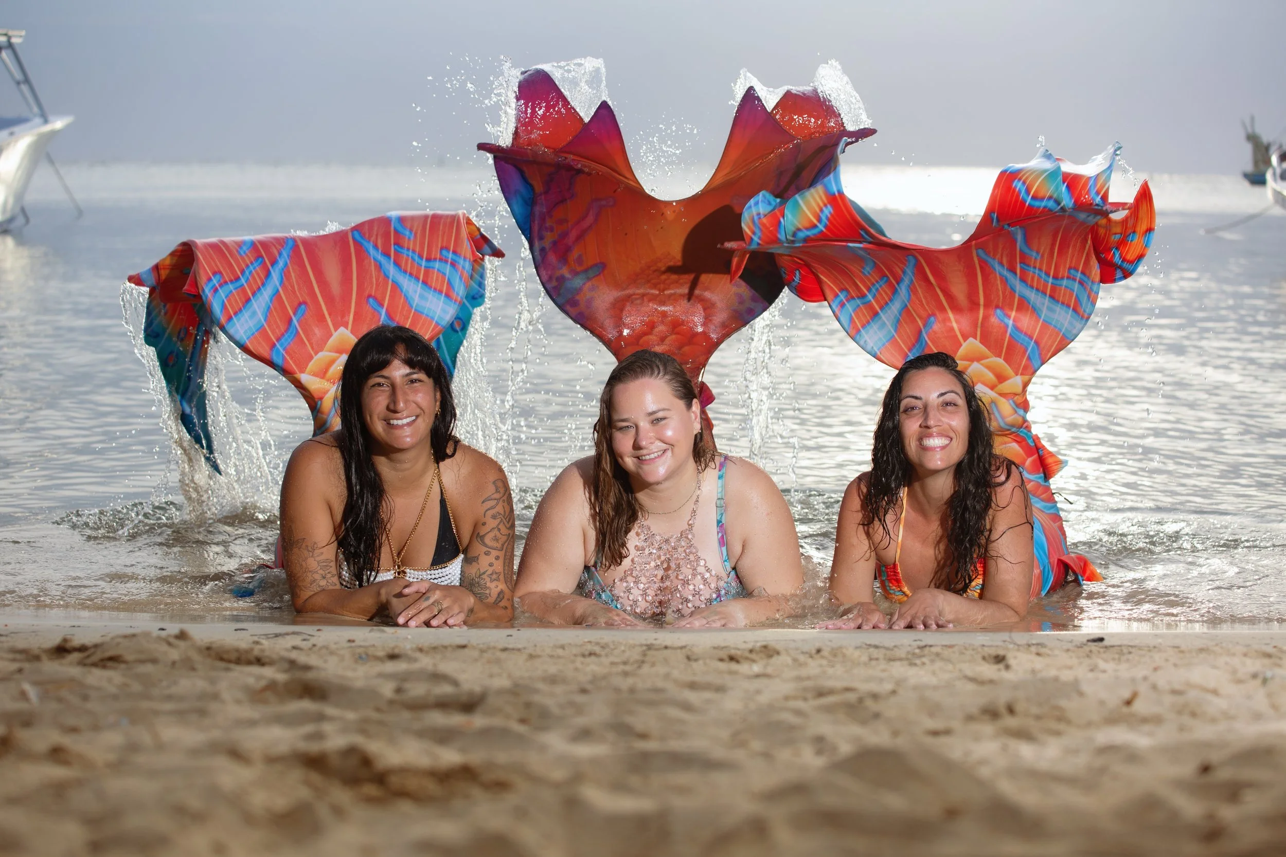 Three women lying on the beach with colorful mermaid tail floaties on their backs, splashing water, with a boat in the background.
