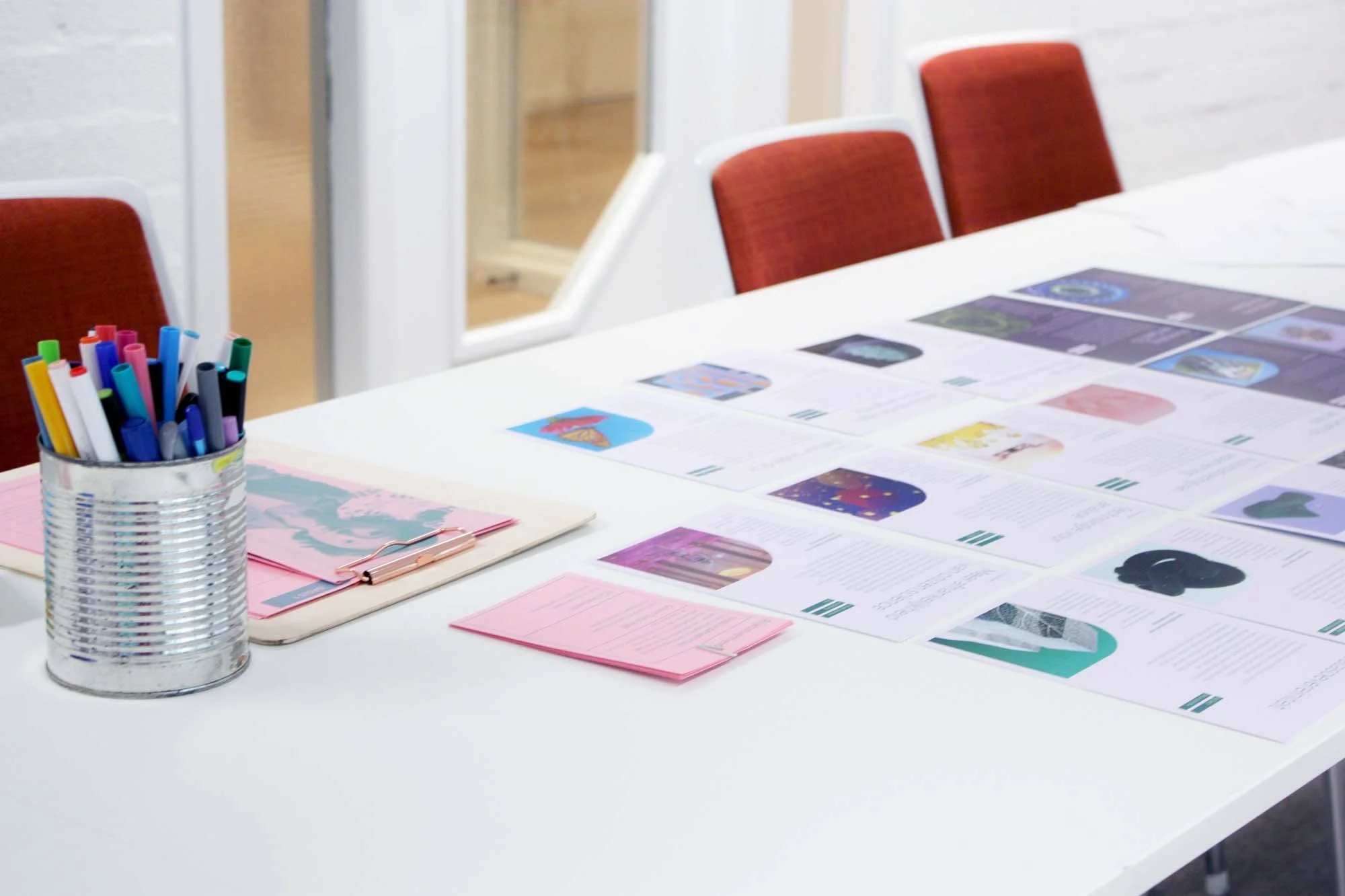 Een wit bureau met verschillende papierwerk en een emmer met gekleurde stiften. Er zijn rode stoelen op de achtergrond en een raam met een deur.