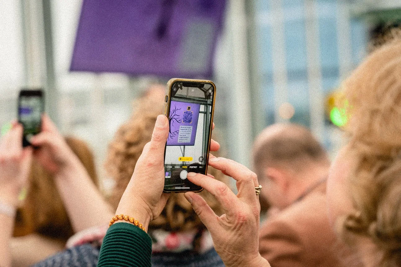 Een persoon die een foto maakt van een bord met een QR-code en tekst, terwijl meerdere mensen in de achtergrond aanwezig zijn.