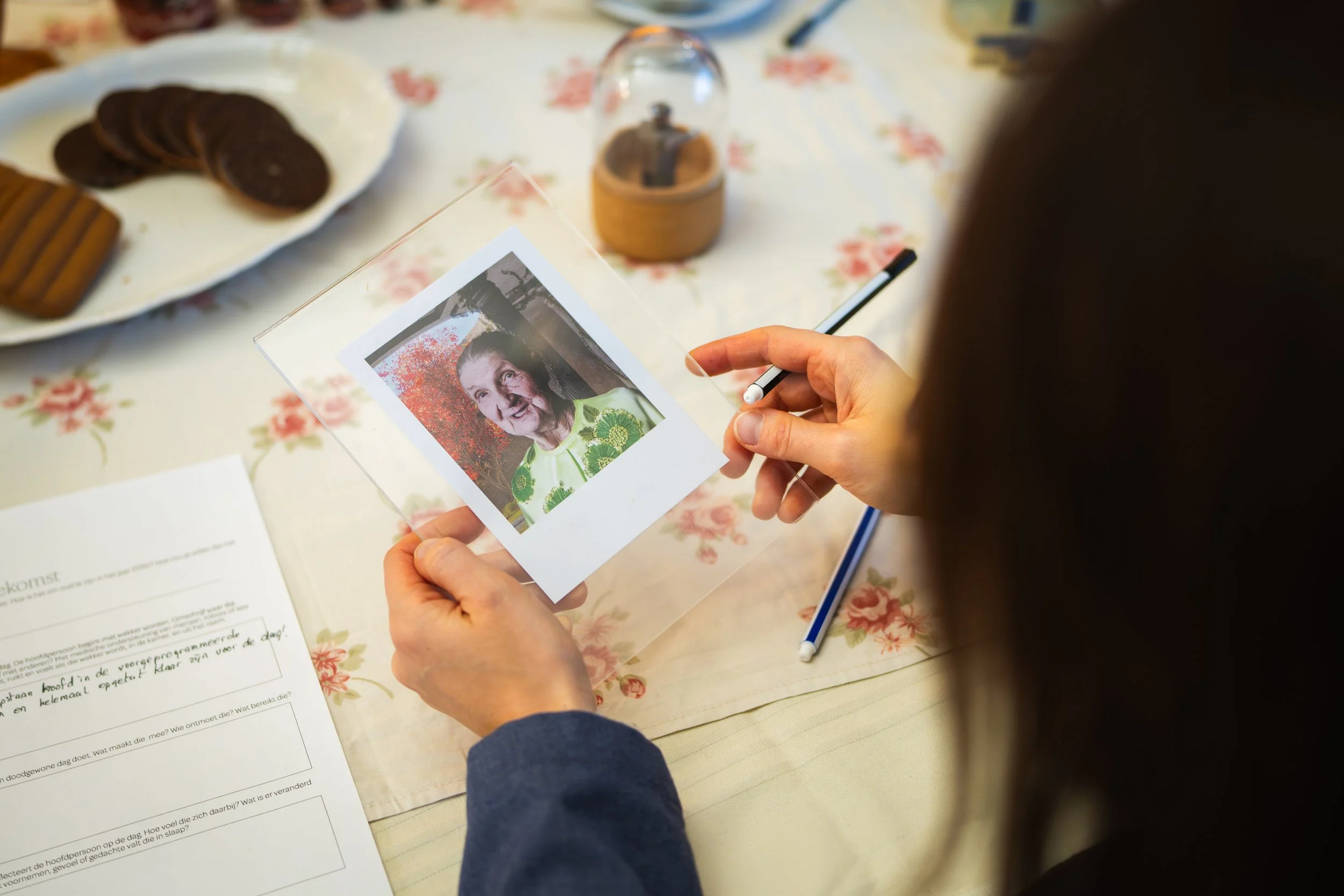 Persoon houdt een foto vast van een oudere vrouw met een groene blouse, sitend aan een tafel met een pen en papier, een bord met koekjes op de achtergrond.