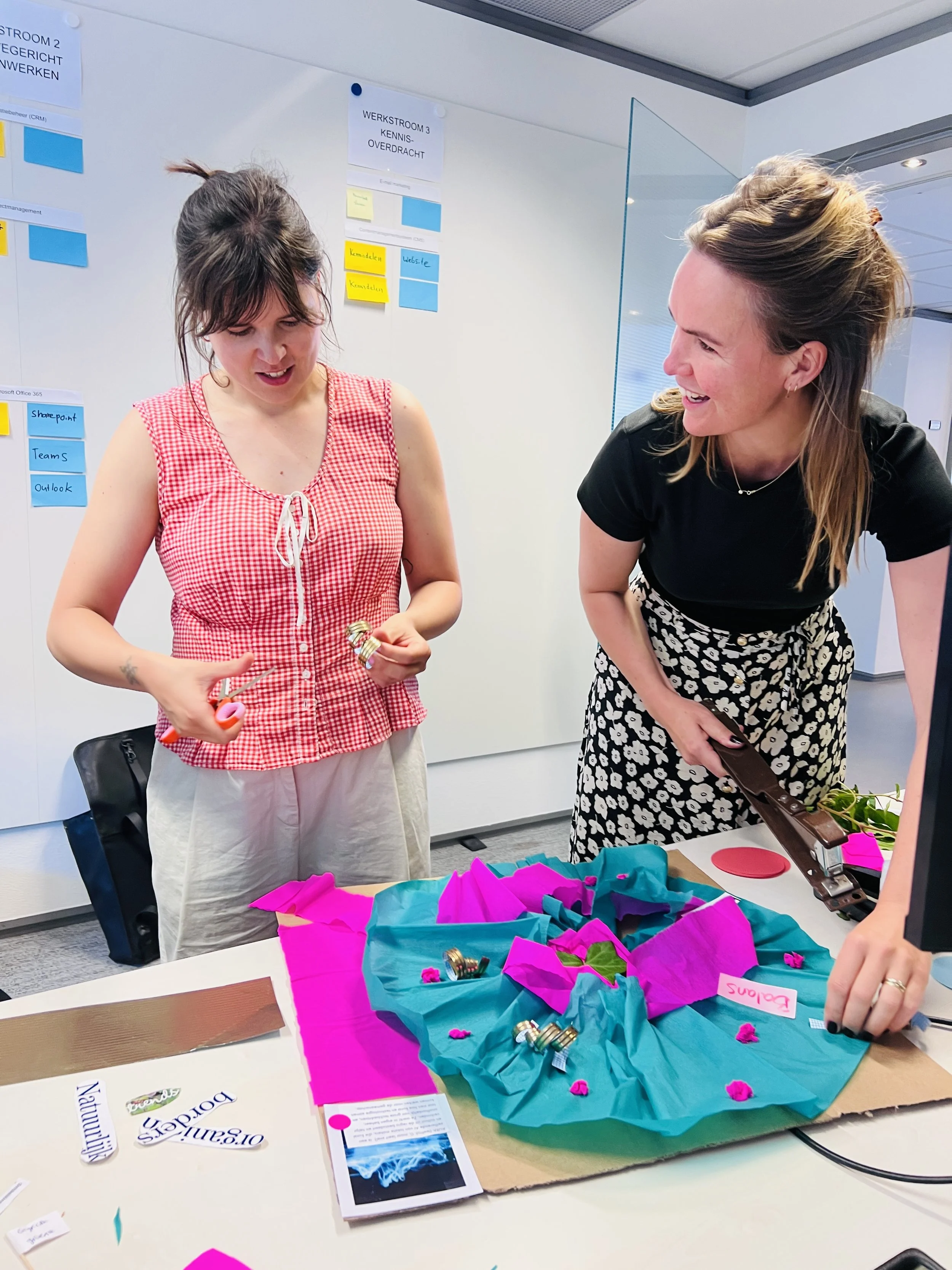 Twee vrouwen lachen en kijken naar een kleurrijk kunstproject met roze en blauwe papieren en kleine voorwerpen op een tafel.