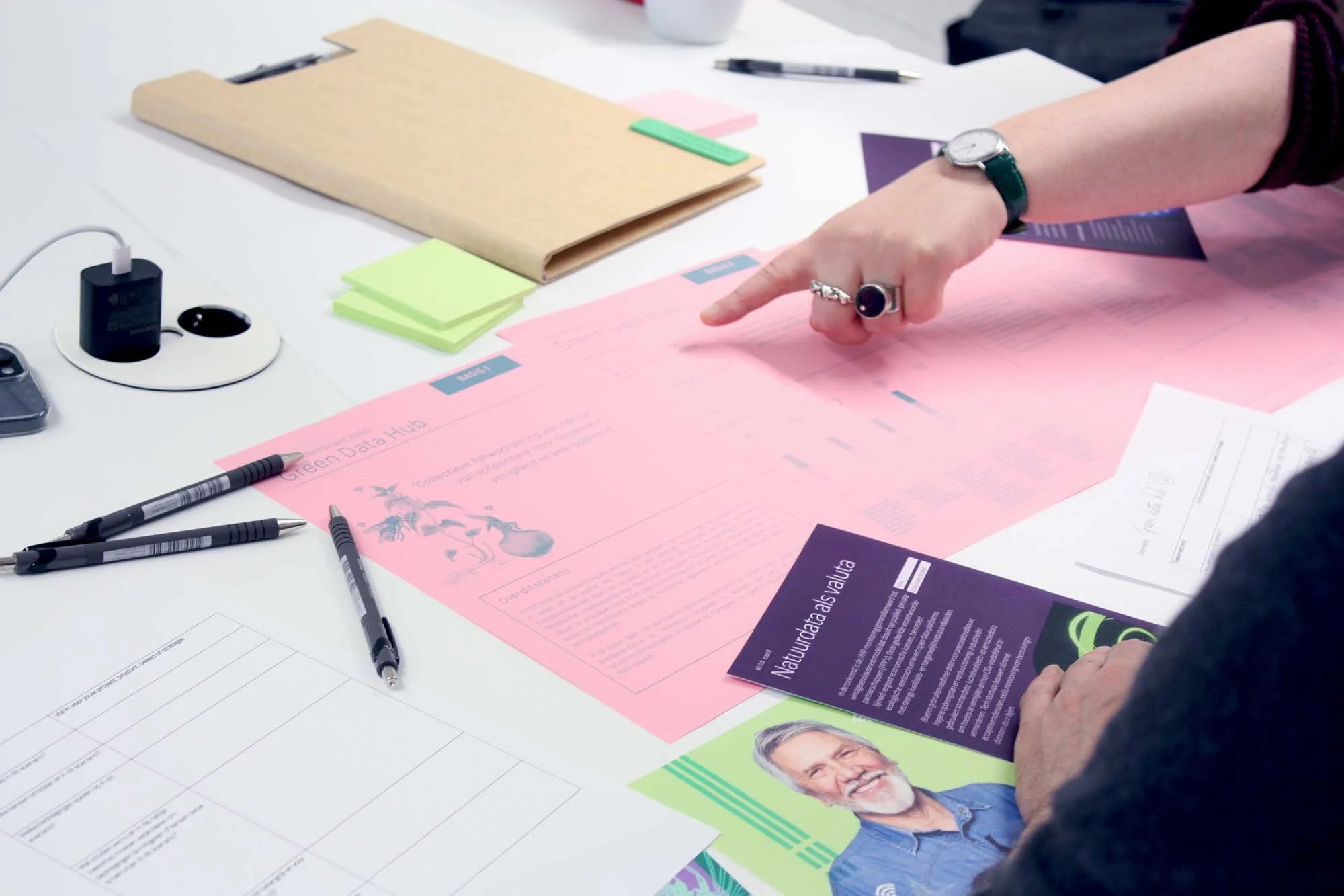 Persoon die wijst naar een roze ingezoomd document op een tafel met diverse documenten, pennen, notitieblok en andere kantoorbenodigdheden.