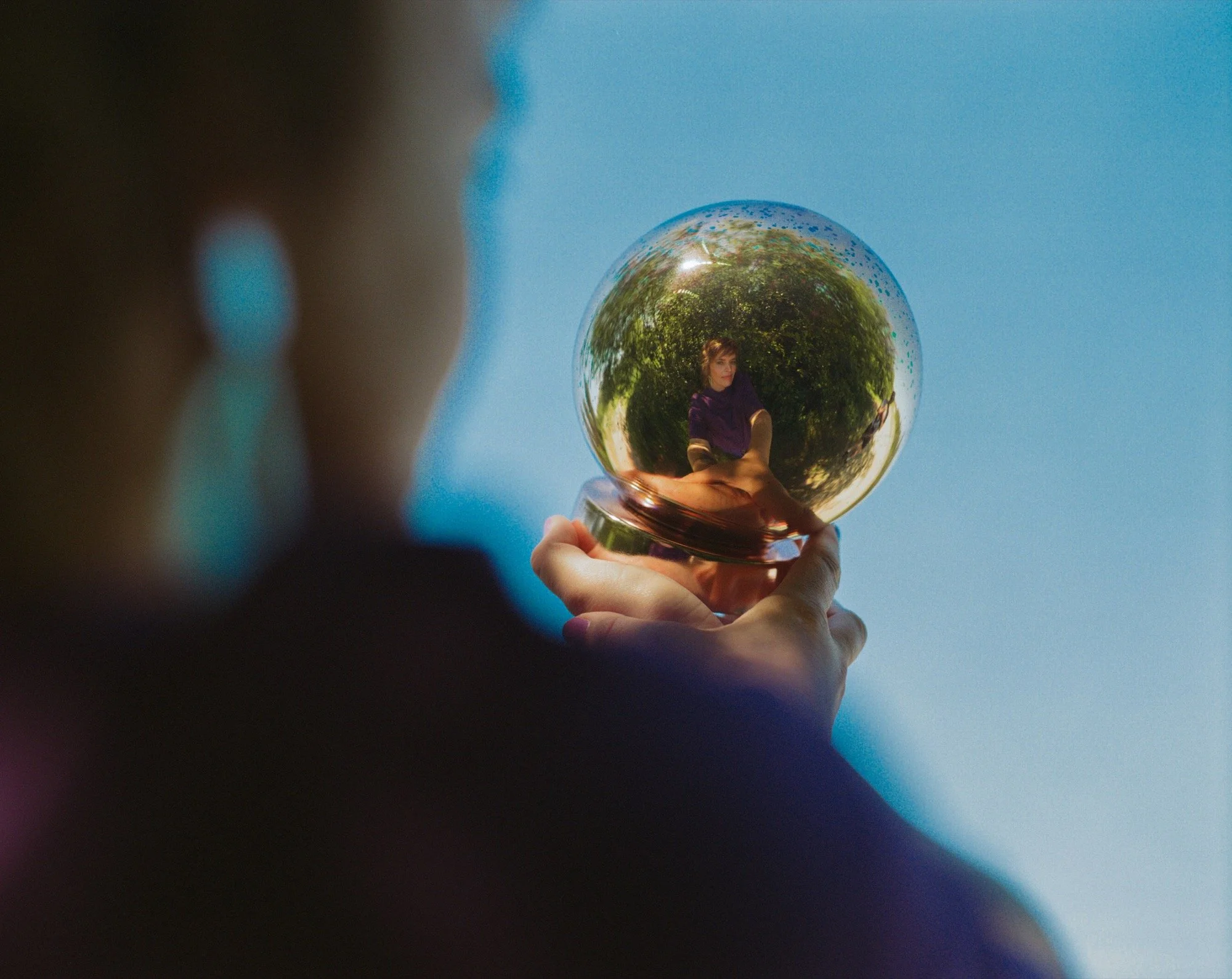 Persoon houdt een glazen bol vast met een reflectie van een vrouw en groene bomen erin.