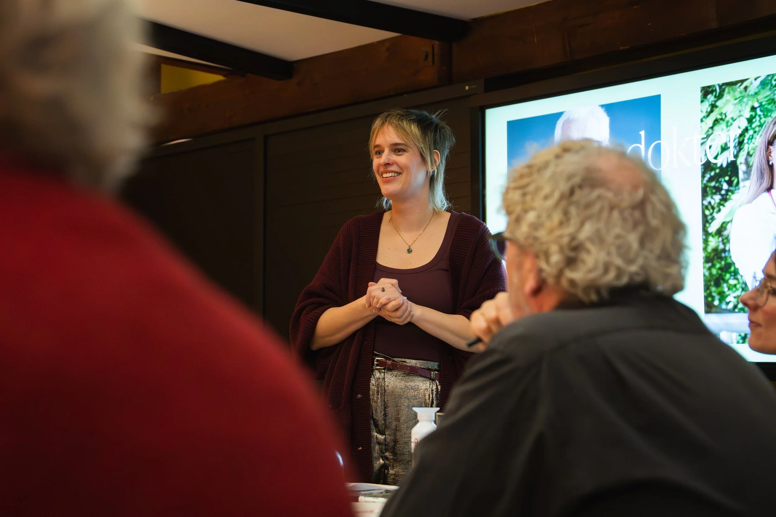Vrouw geeft presentatie aan een groep mensen in een vergaderzaal, achter haar een scherm met een afbeelding.
