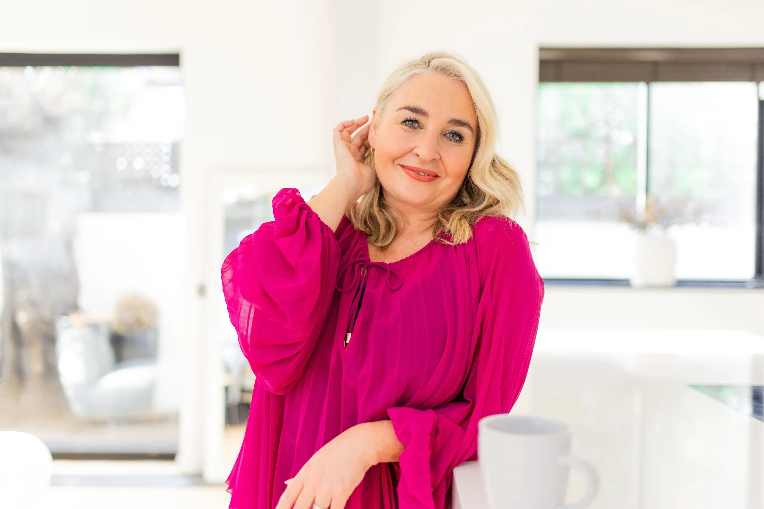 Blonde woman in vibrant magenta blouse leaning against kitchen island , smiling at the camera while brushing some of her hair behind her ear.