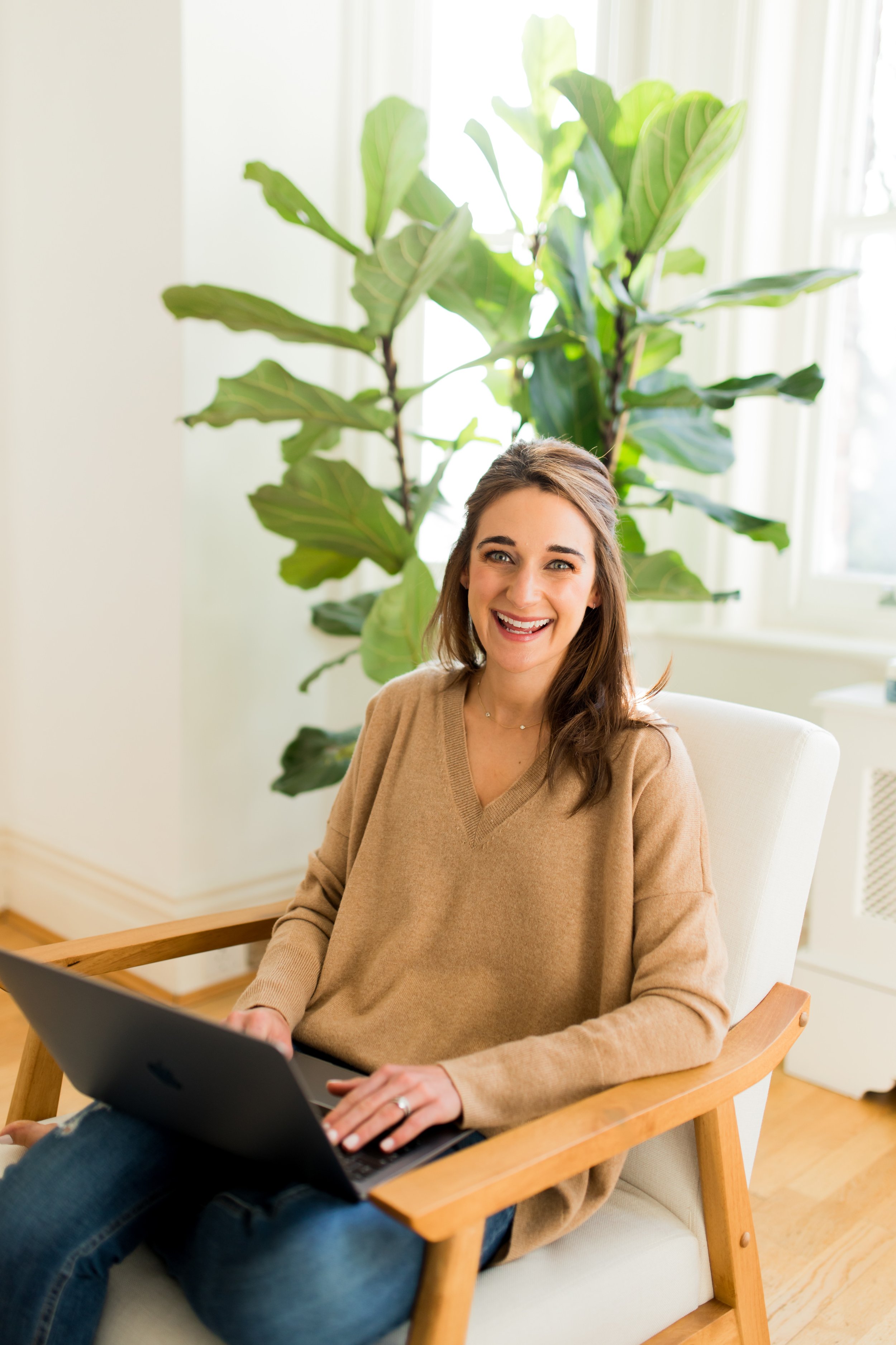 woman with long brown hair in a beige/light brown seater sitting in a lounge chair with her laptop. she's looking at the camera, smiling.