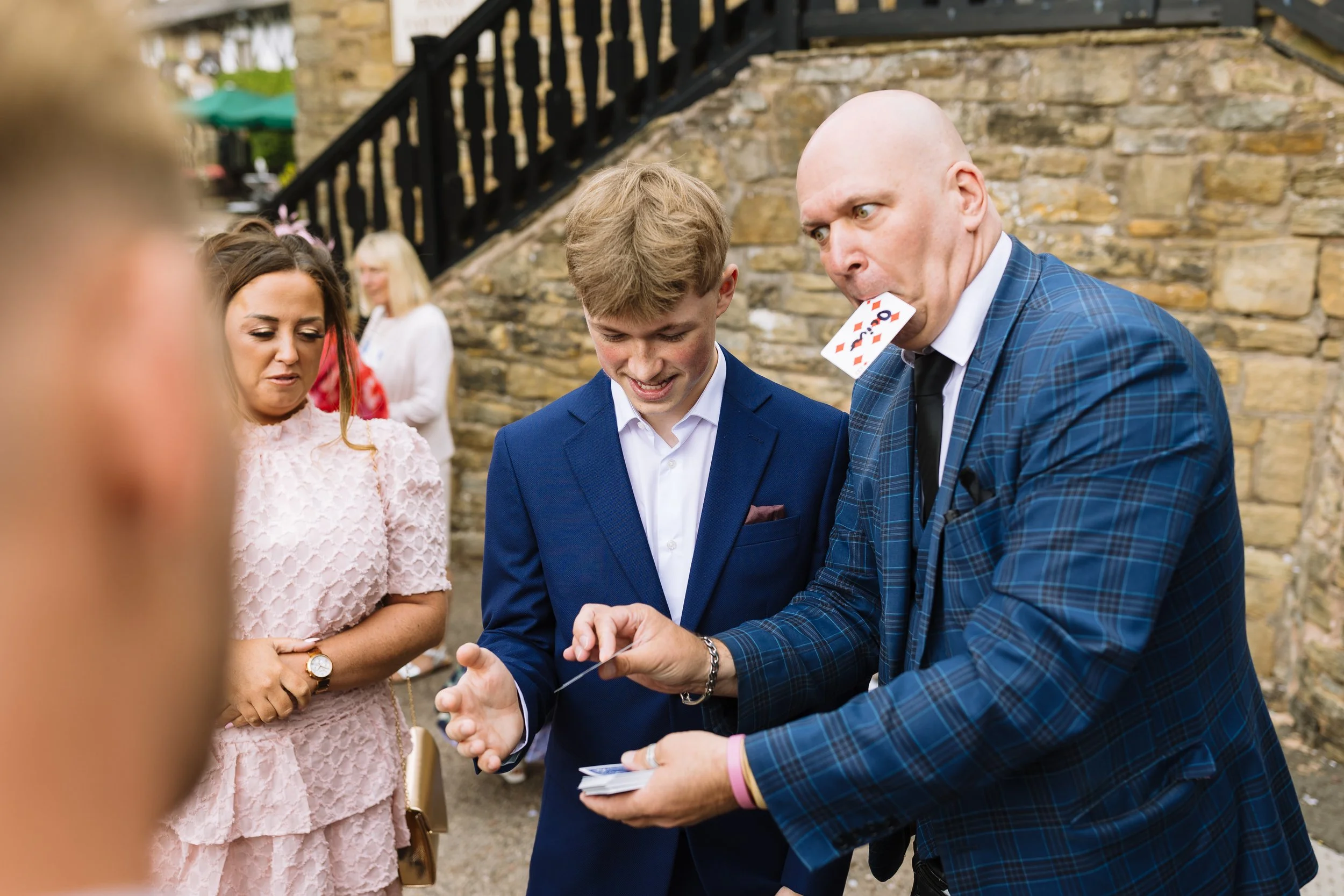 A group of people at an outdoor event with a man performing a magic trick with a card while a woman in a pink dress and a young man in a blue suit watch.