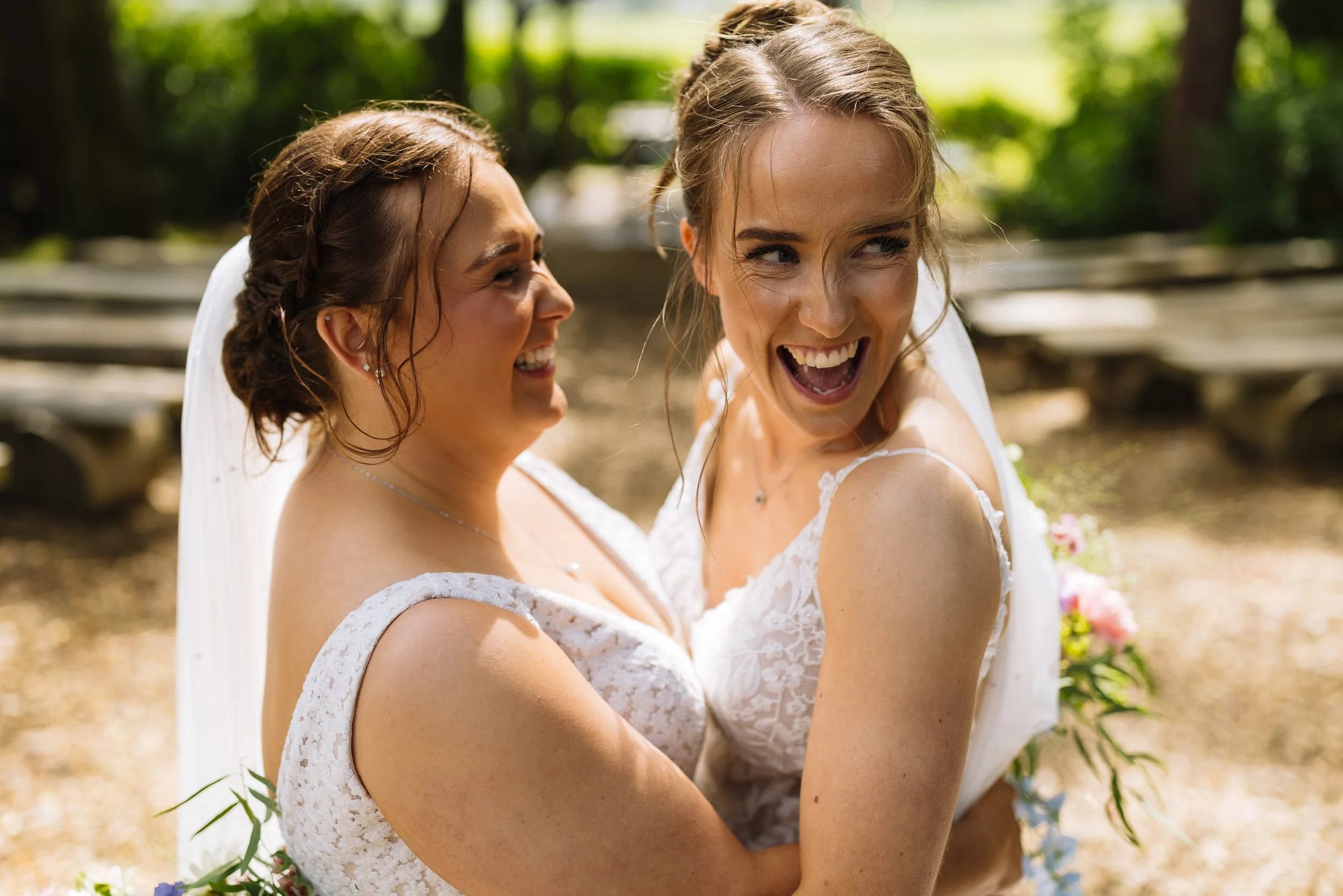 Two women in wedding dresses hugging and laughing outdoors on a sunny day.