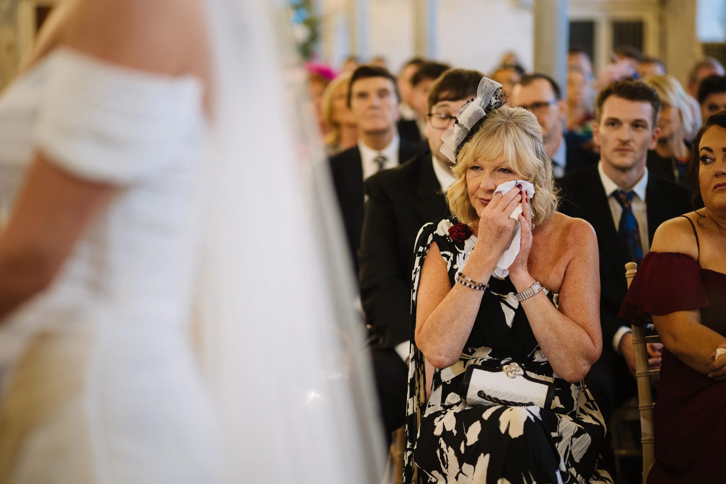 A woman in a floral black and white dress sitting in a formal event, wiping away tears with a tissue, with other seated guests in formal attire visible in the background.
